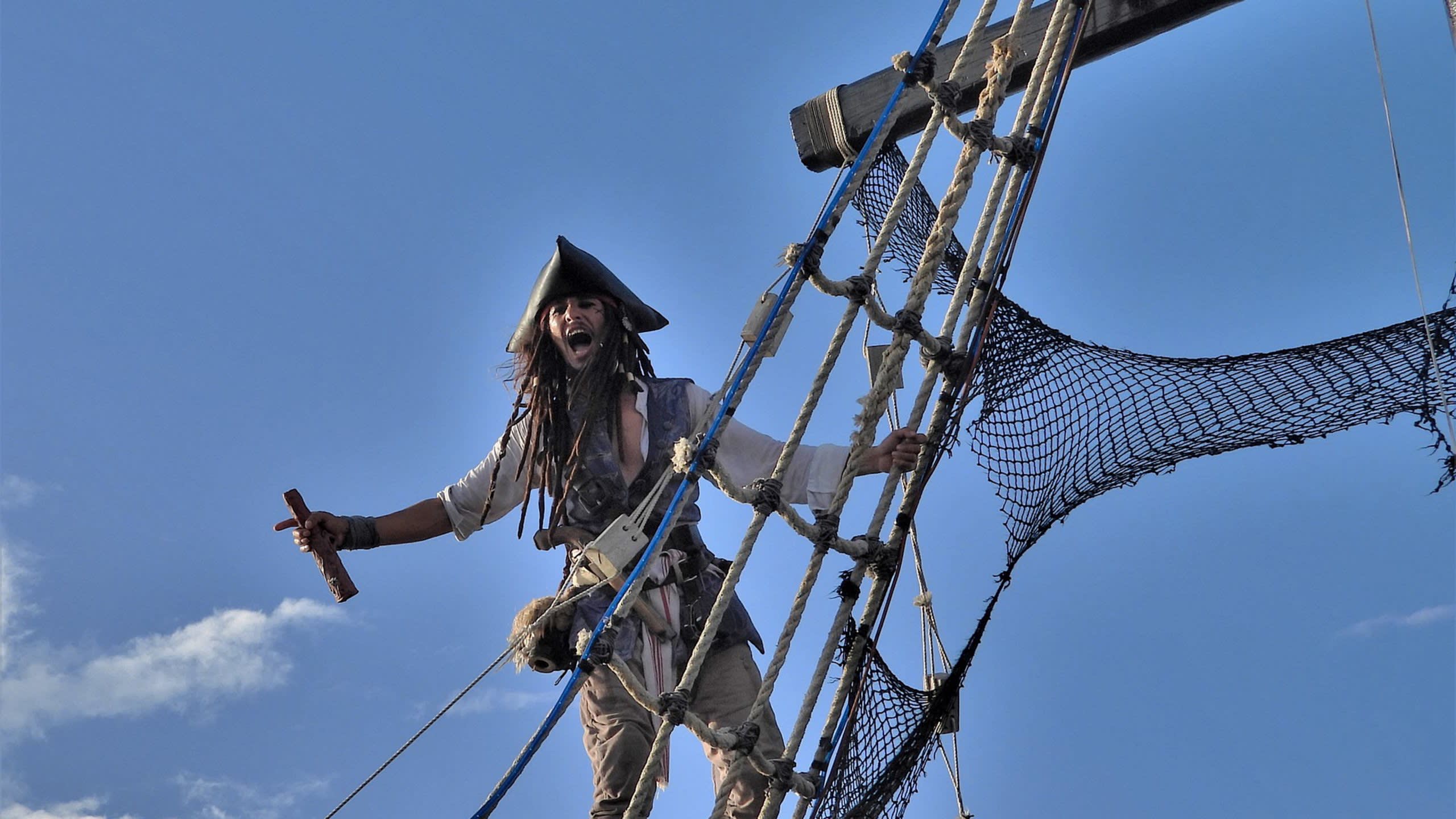 A pirate with dreadlocks and a tricorn hat yells while climbing a ship's rigging against a bright blue sky.