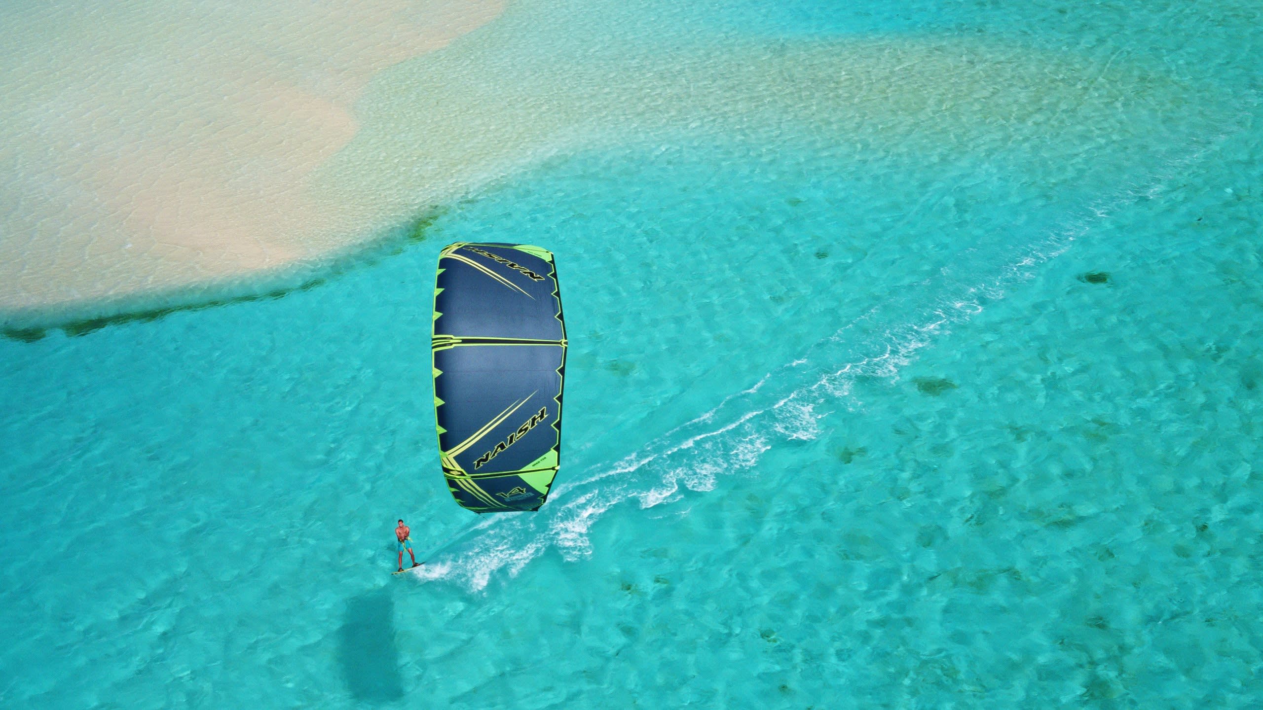 A lone kitesurfer glides across crystal-clear turquoise waters near a sandy shore.
