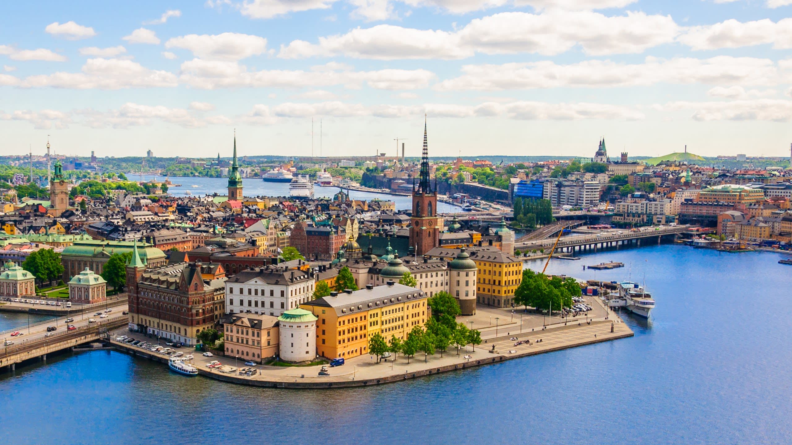 Stockholm's historic waterfront buildings and bustling waterways create a picturesque urban landscape under a bright, cloudy sky.