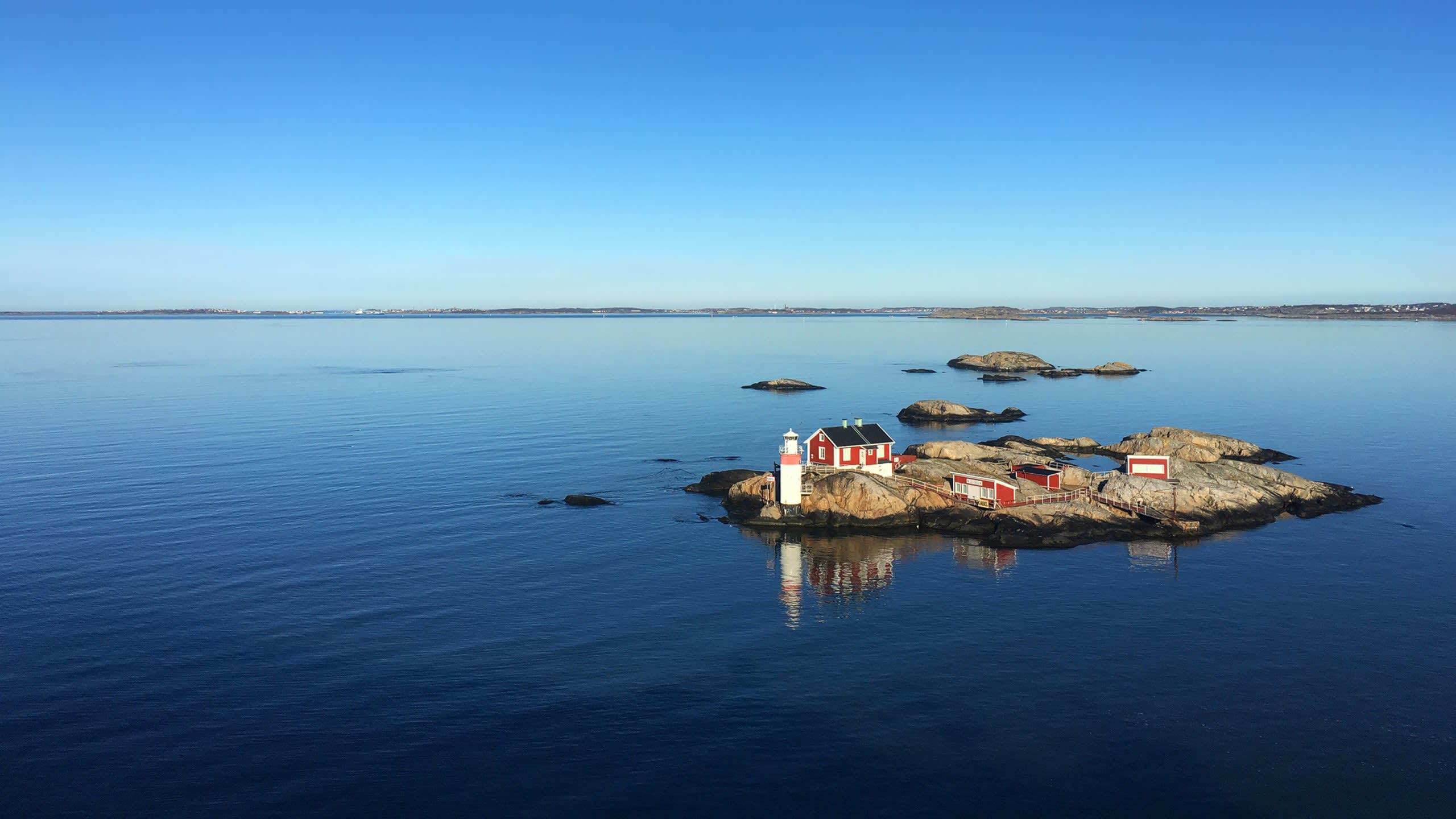 A charming red lighthouse and accompanying buildings stand proudly on a rocky island surrounded by calm, blue waters under a clear sky.