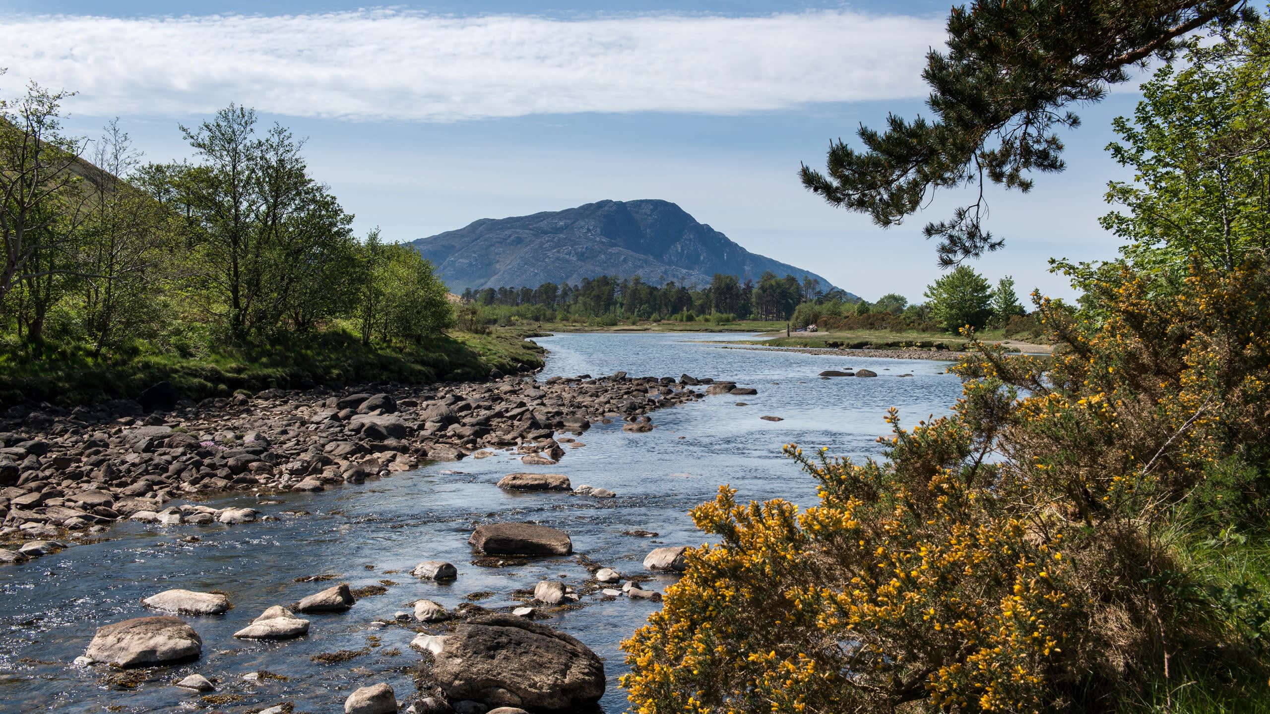 A wide river flows through a rocky landscape towards a majestic mountain under a partly cloudy sky.