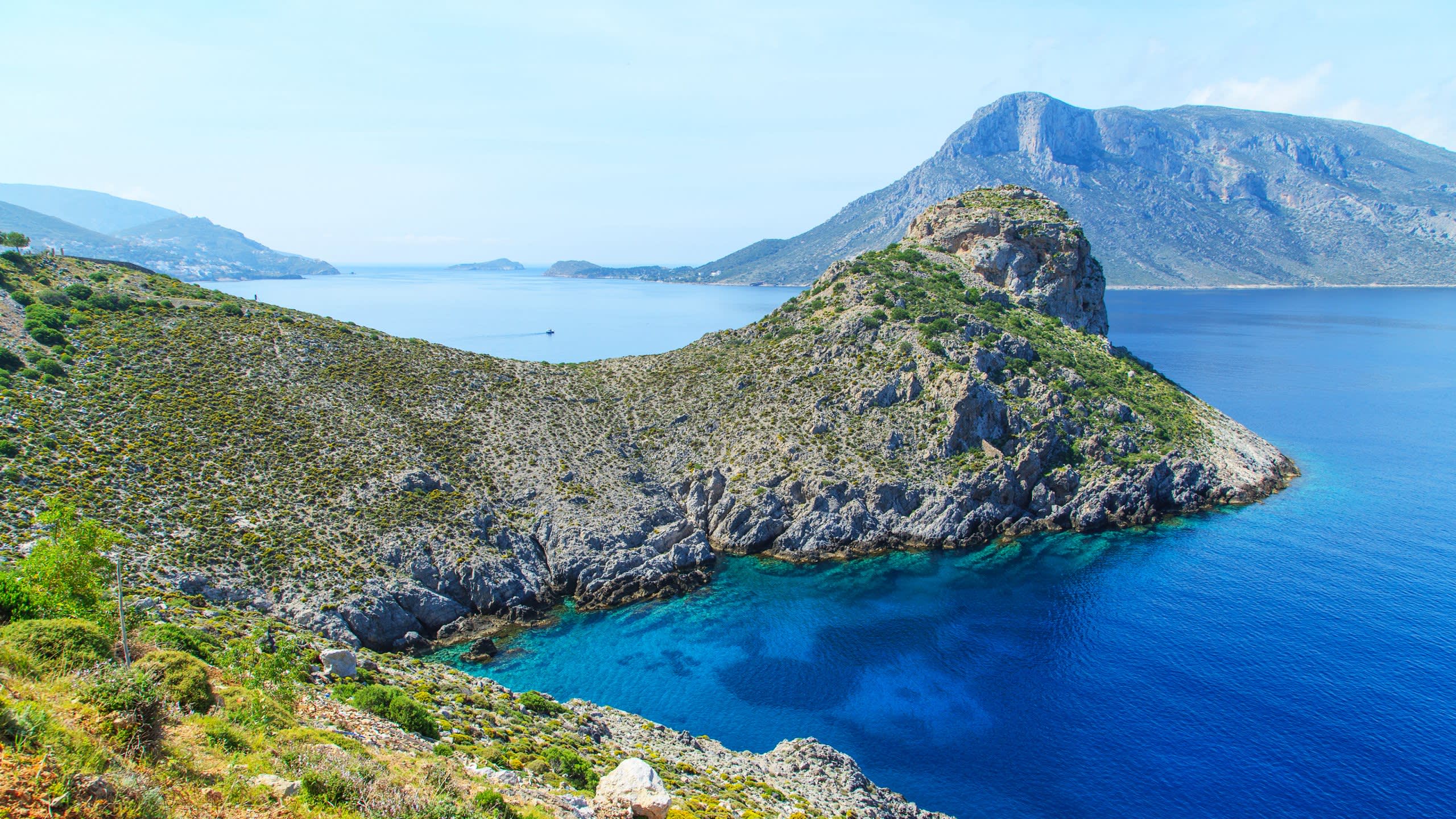 A rugged, green-covered island juts into the crystal-clear turquoise waters of the Aegean Sea under a bright blue sky.