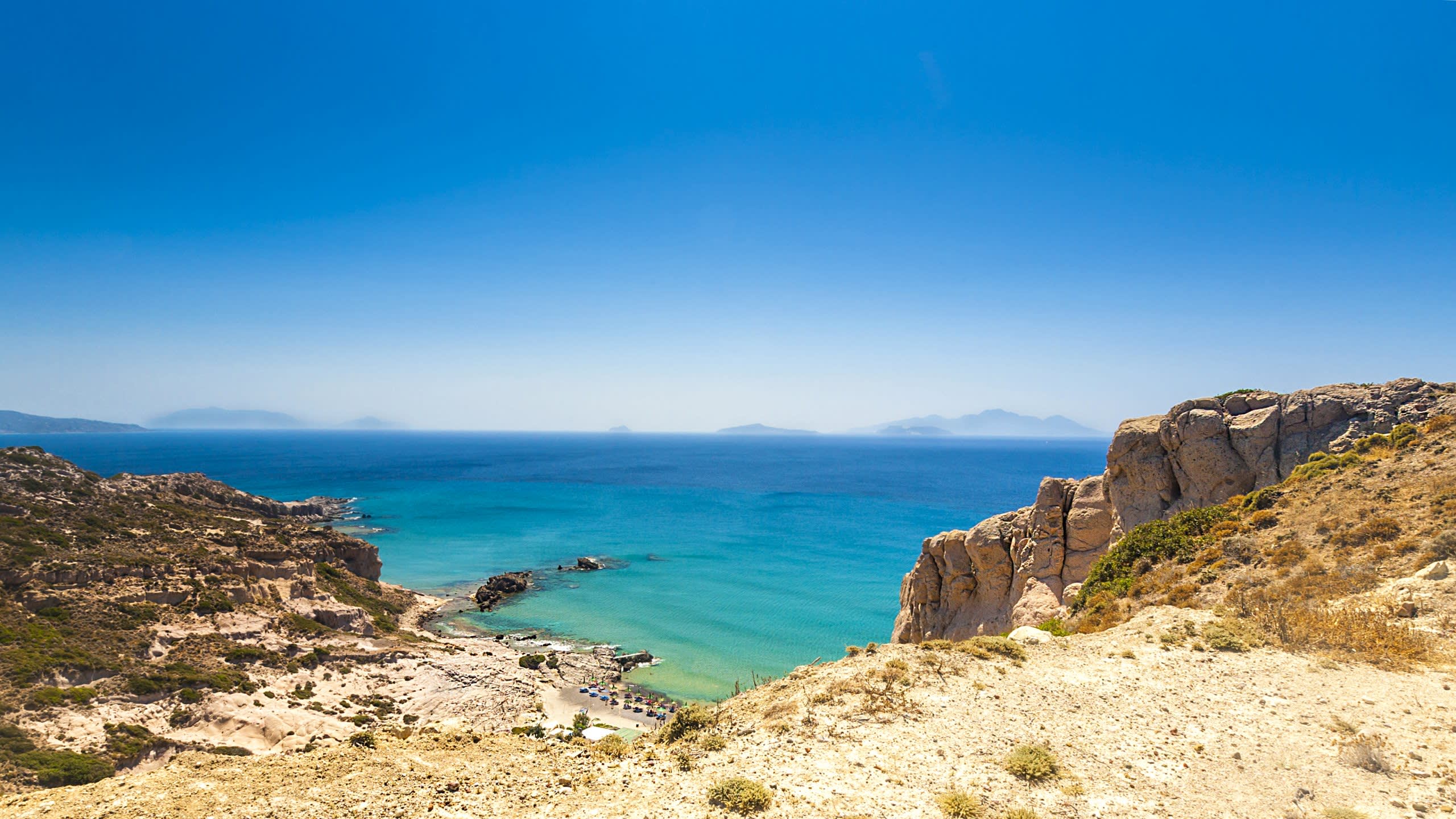 A bright blue sky stretches over a rugged coastline with turquoise waters lapping at a small beach.