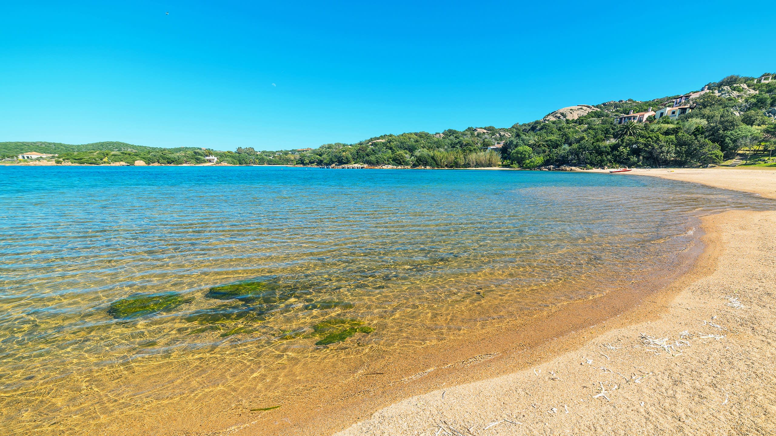 Crystal-clear turquoise waters gently lap a golden sandy beach nestled beneath lush green hills dotted with elegant villas.