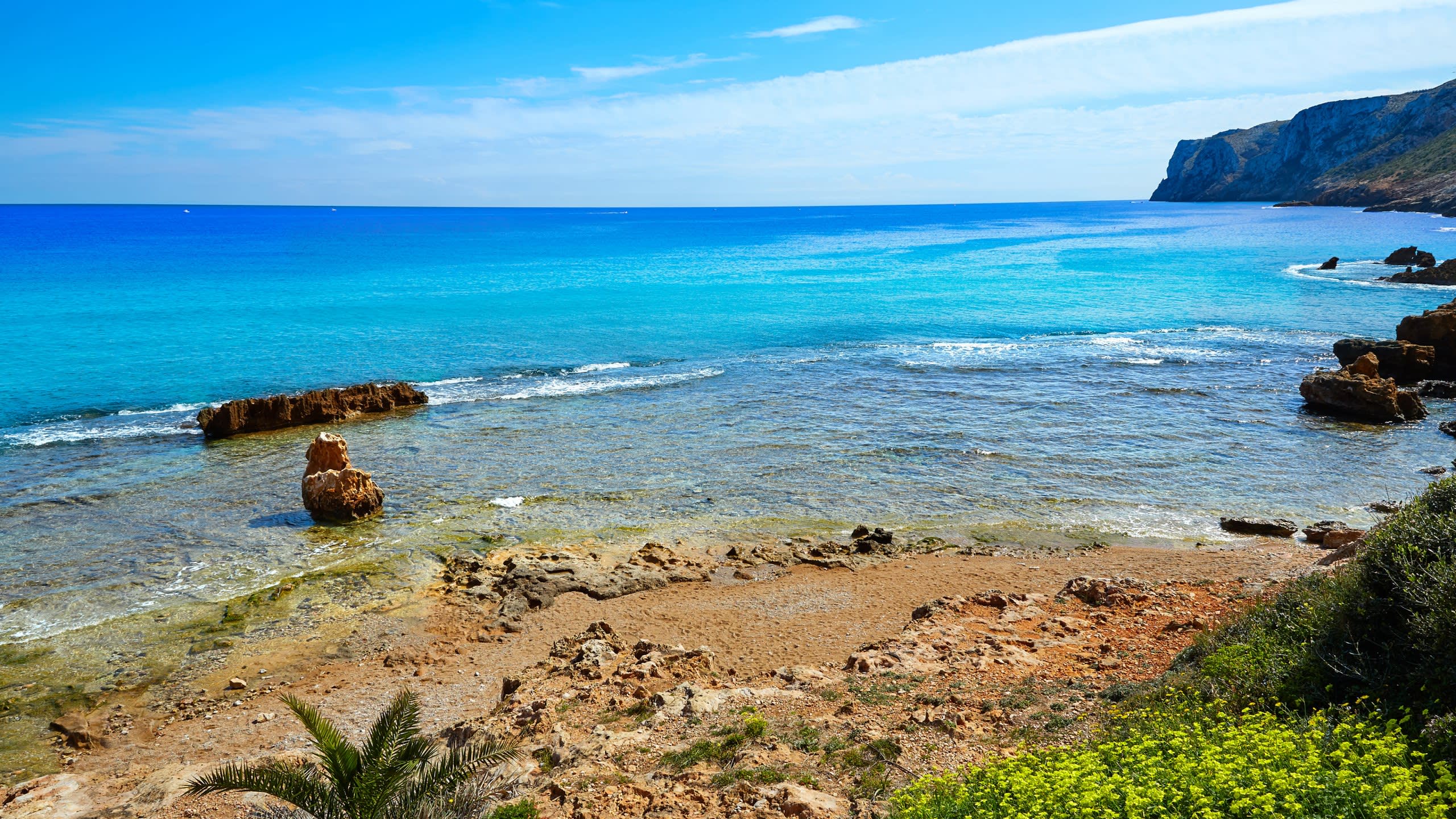 The turquoise waters of a secluded cove gently lap against a sandy beach dotted with rugged rocks and lush green vegetation.