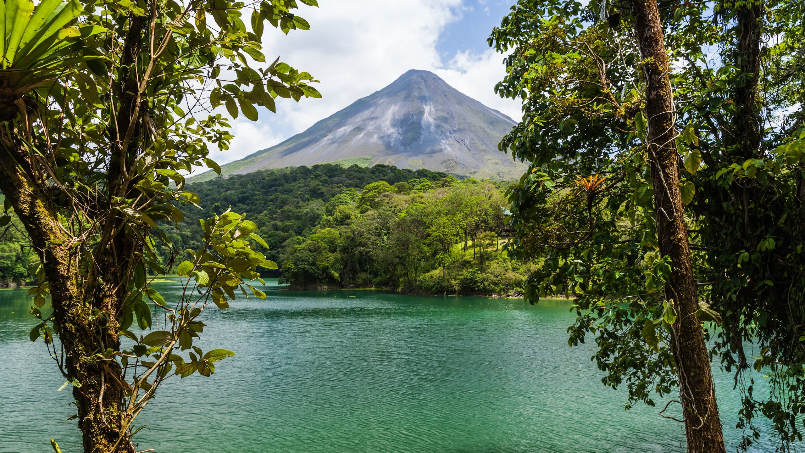 A majestic volcano looms over a tranquil emerald lake, framed by lush tropical foliage.