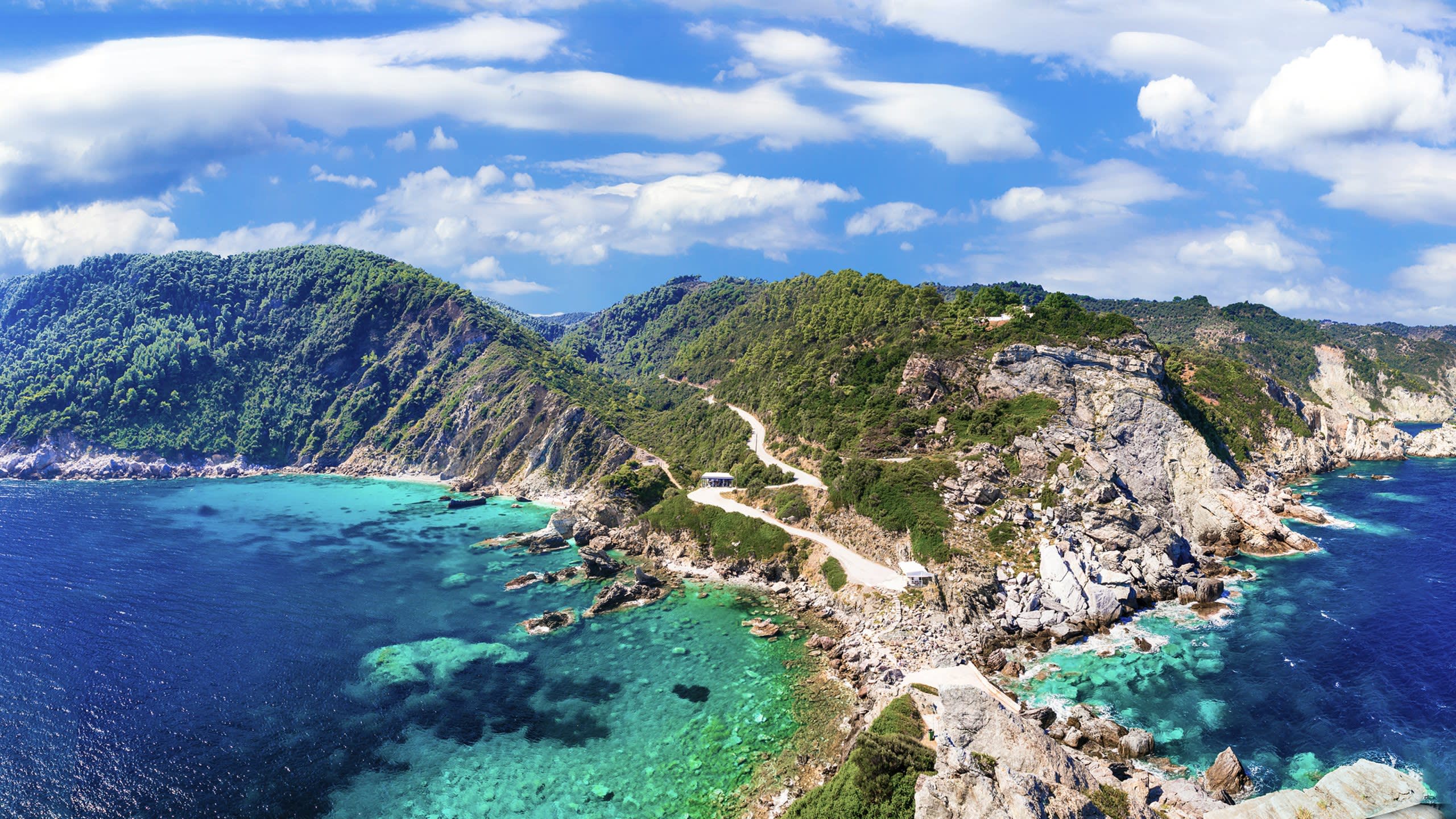 A winding road hugs the lush green mountainside overlooking the crystal-clear turquoise waters of a Mediterranean coastline.
