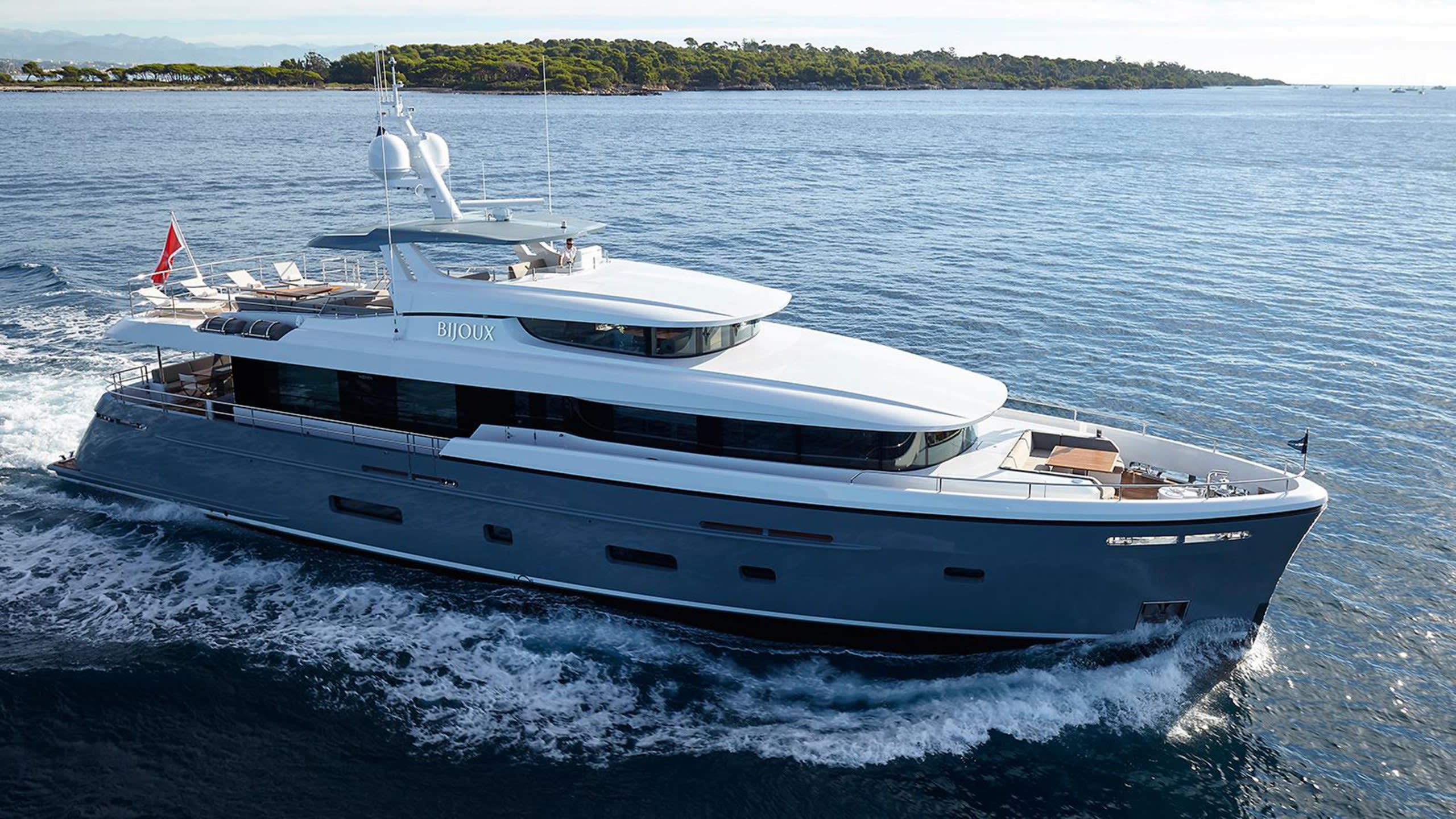 A sleek grey and white yacht named Bijoux cuts through the blue waters off a tree-covered coastline.