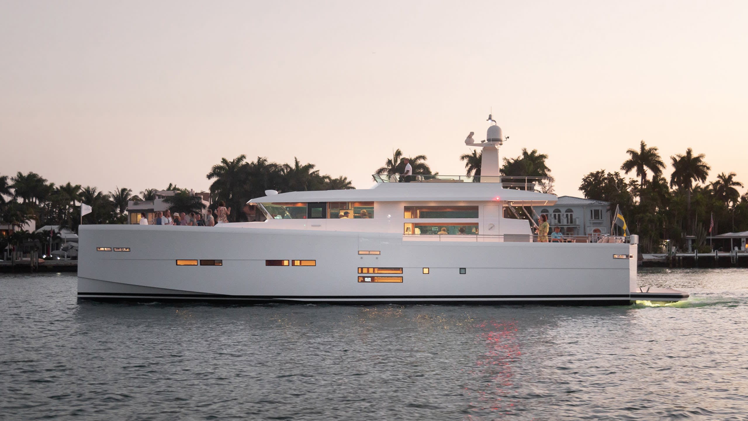 A sleek white yacht glides through calm waters at dusk, its windows illuminated against a pastel sky.