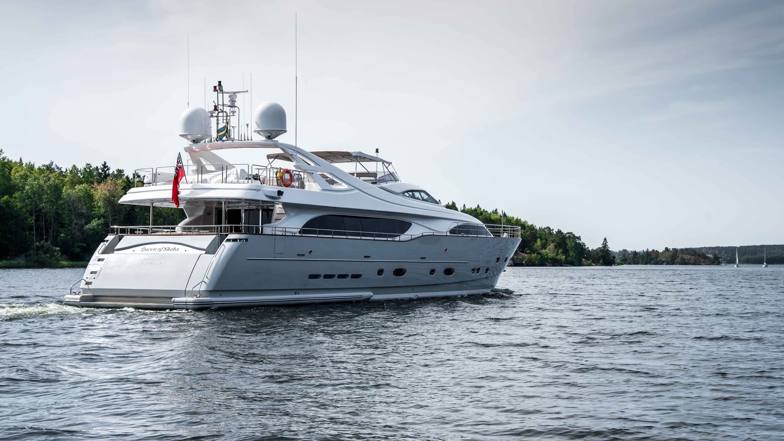 The elegant yacht "Queen of Sheba" cruises through calm waters near a tree-lined shore.