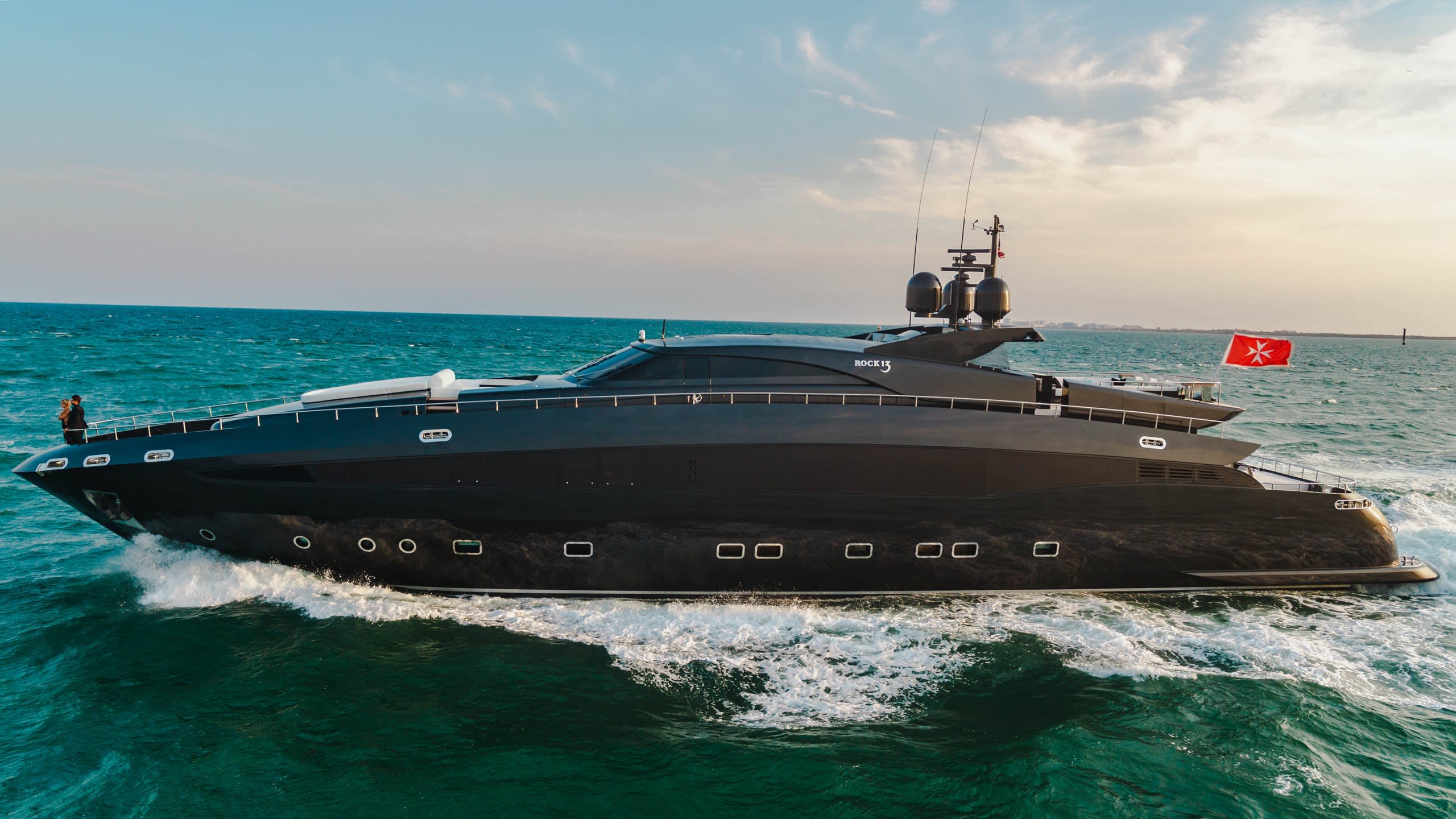 A sleek black yacht named "ROCK13" cuts through the turquoise ocean under a soft, hazy sky.
