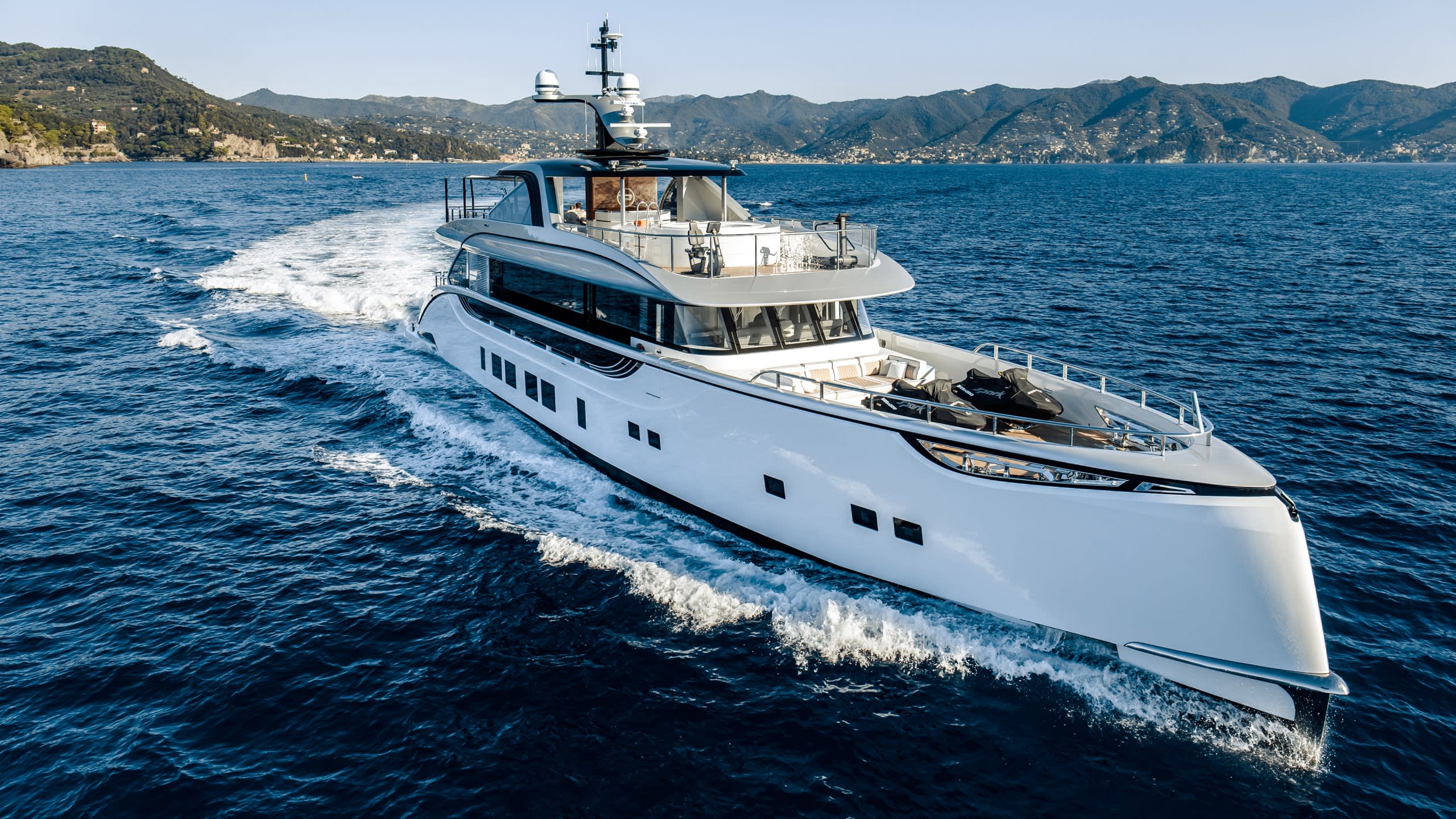 A large, luxurious white yacht is sailing across the deep blue waters, with a mountainous coastal landscape visible in the background.