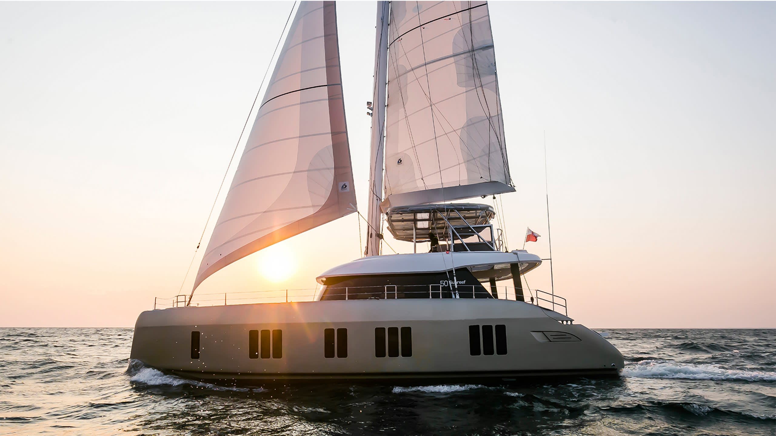 A sleek catamaran glides through calm waters at sunset, its sails catching the golden light.