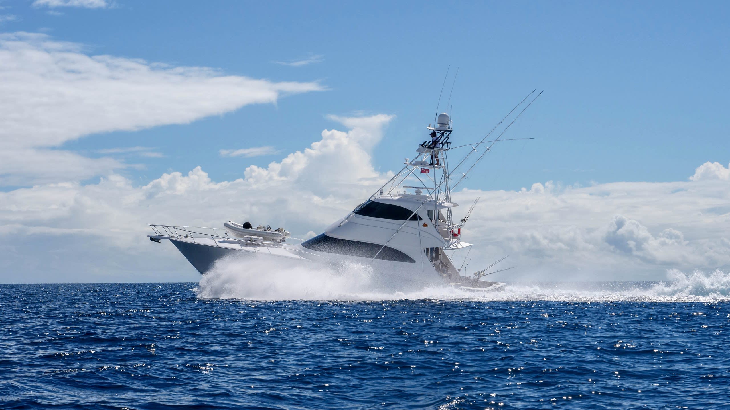 A powerful sportfishing yacht cuts through the deep blue ocean, kicking up a dramatic spray under a bright, cloudy sky.