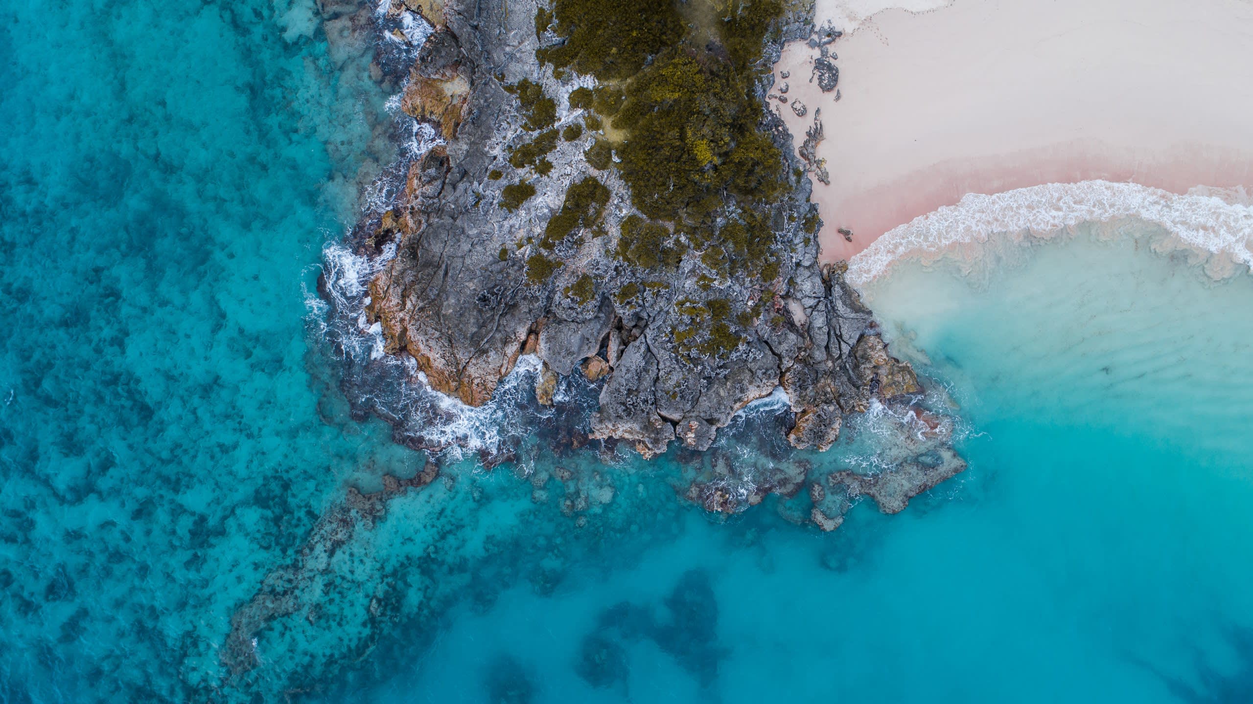 Turquoise waters gently lap against a rugged rocky coastline and a soft pink sand beach.