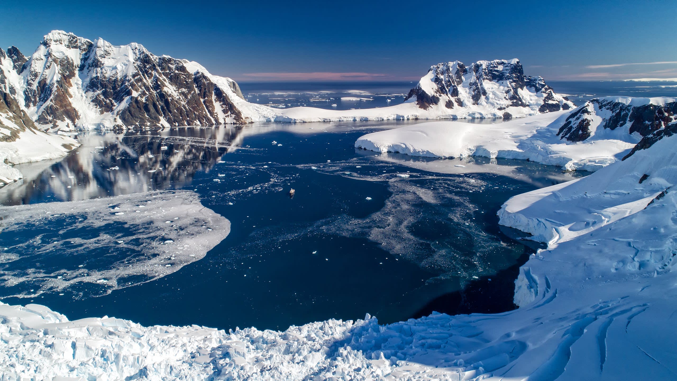 A majestic ship navigates the icy waters of Antarctica, surrounded by towering snow-capped mountains and floating icebergs under a clear blue sky.