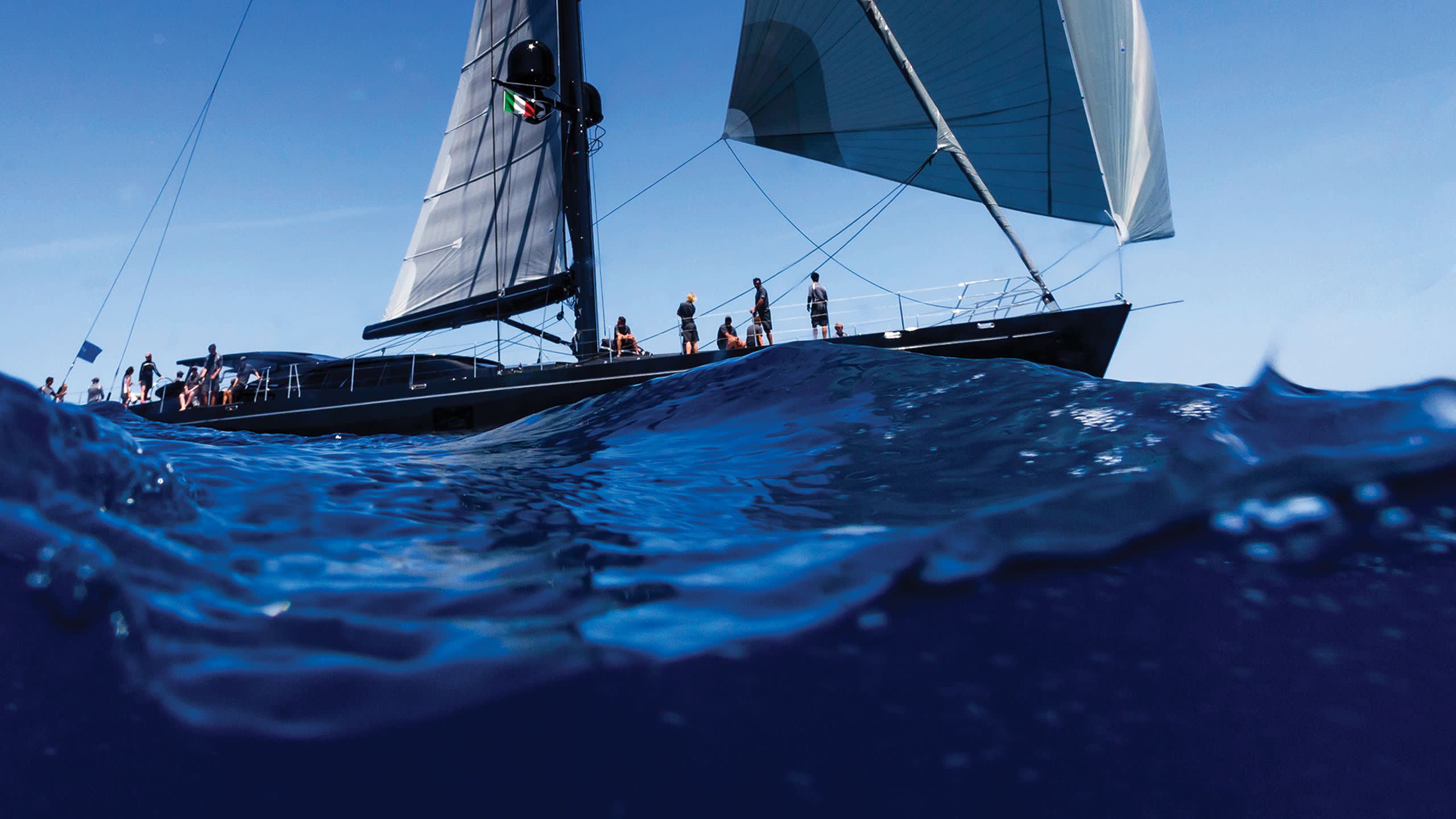 A sleek black sailboat glides through the deep blue ocean under a clear sky.