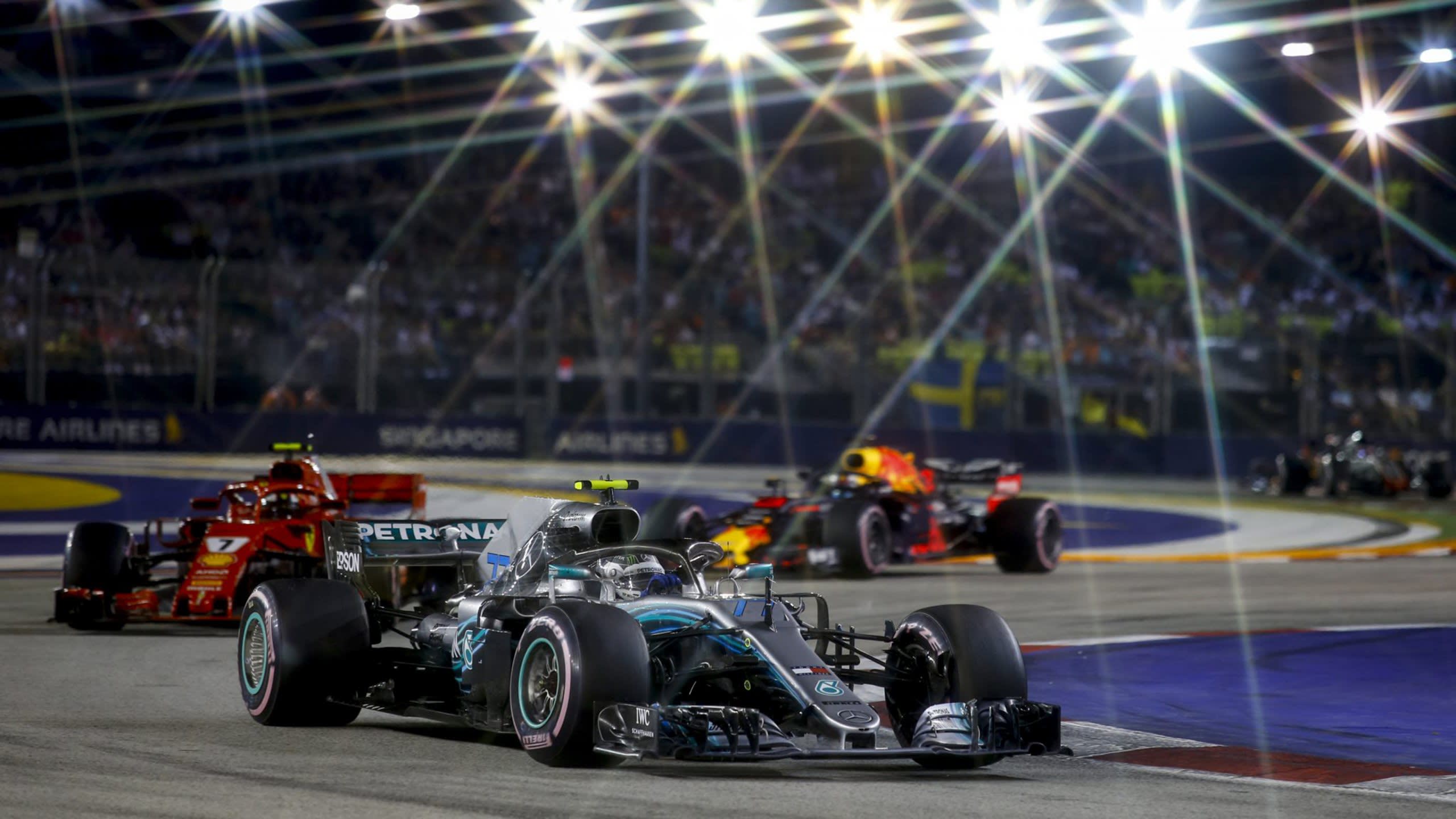 Formula 1 cars race under bright stadium lights on a night circuit, with a packed grandstand in the background.