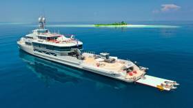 A luxurious yacht named Bold anchors in the clear blue waters near a small tropical island.