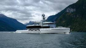 A sleek, modern yacht cruises through calm water with dramatic, cloud-covered mountains rising in the background.