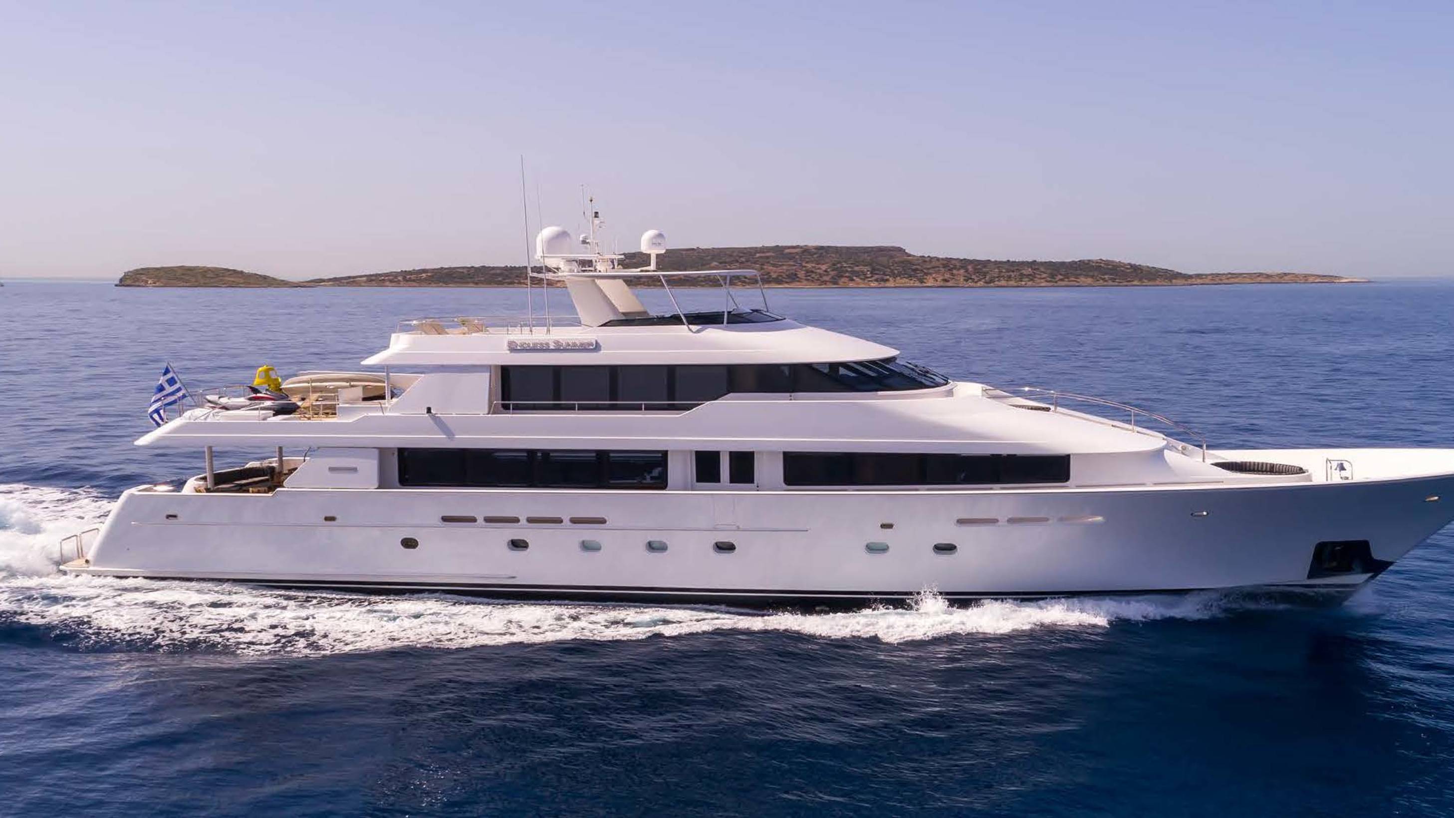 A large white yacht cruises through the deep blue sea, with a distant, rocky island under a clear sky in the background.