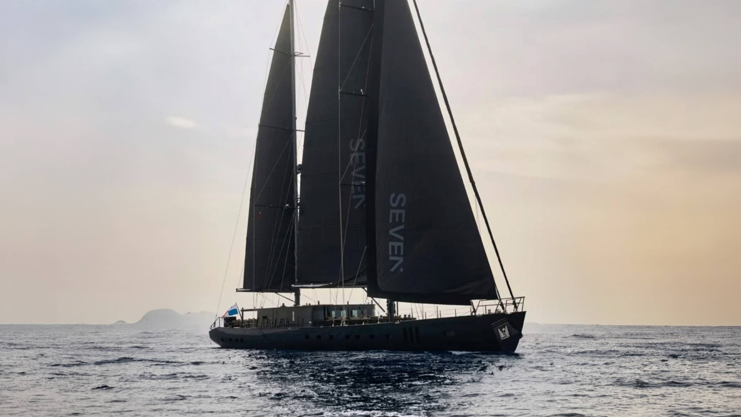 A sleek, black-sailed yacht glides across a calm sea with a hazy, sunlit sky and distant islands in the background.