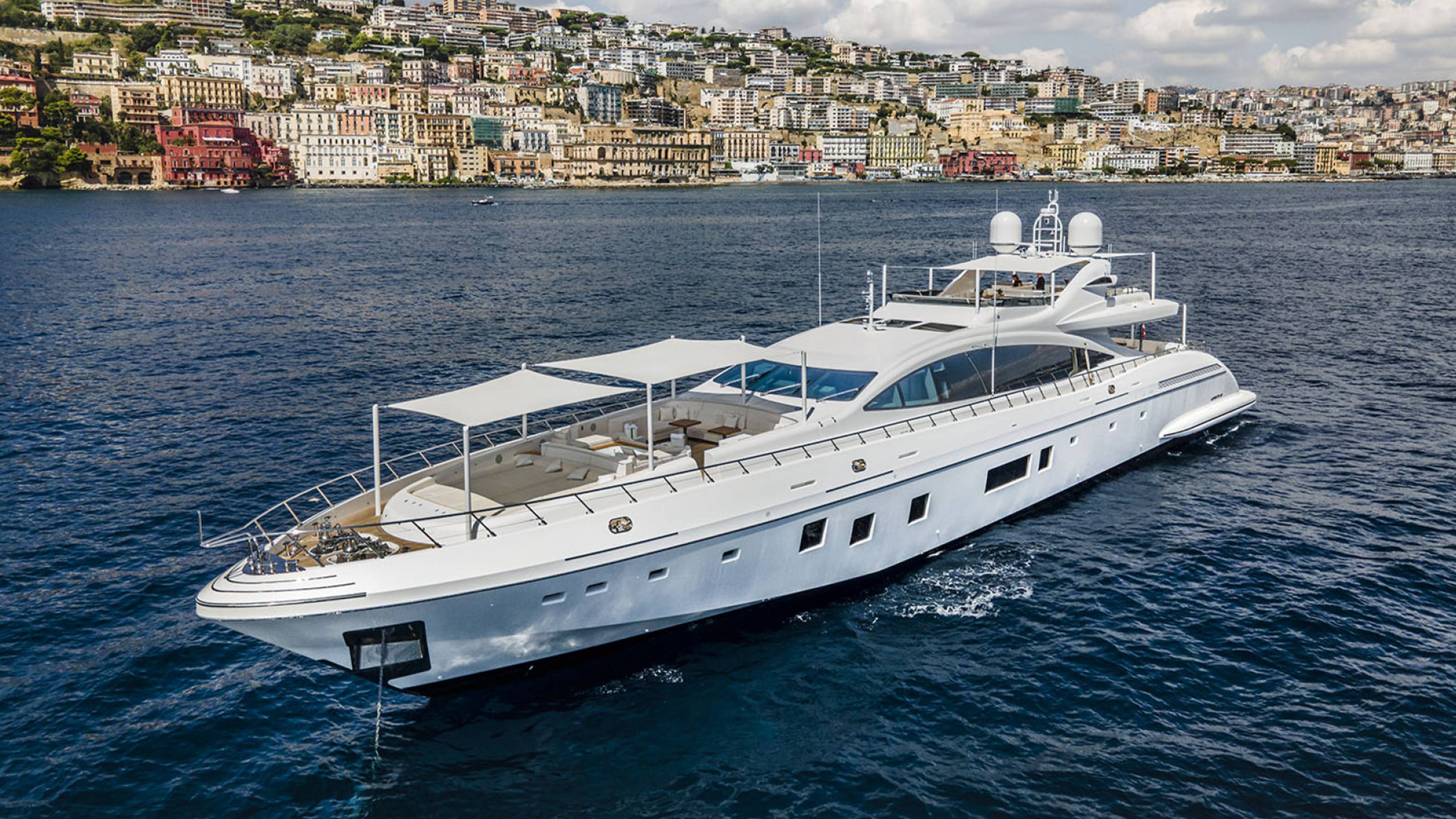 A large, luxurious white yacht is sailing on the deep blue waters of a harbor, with a picturesque city nestled on the hillside in the background.