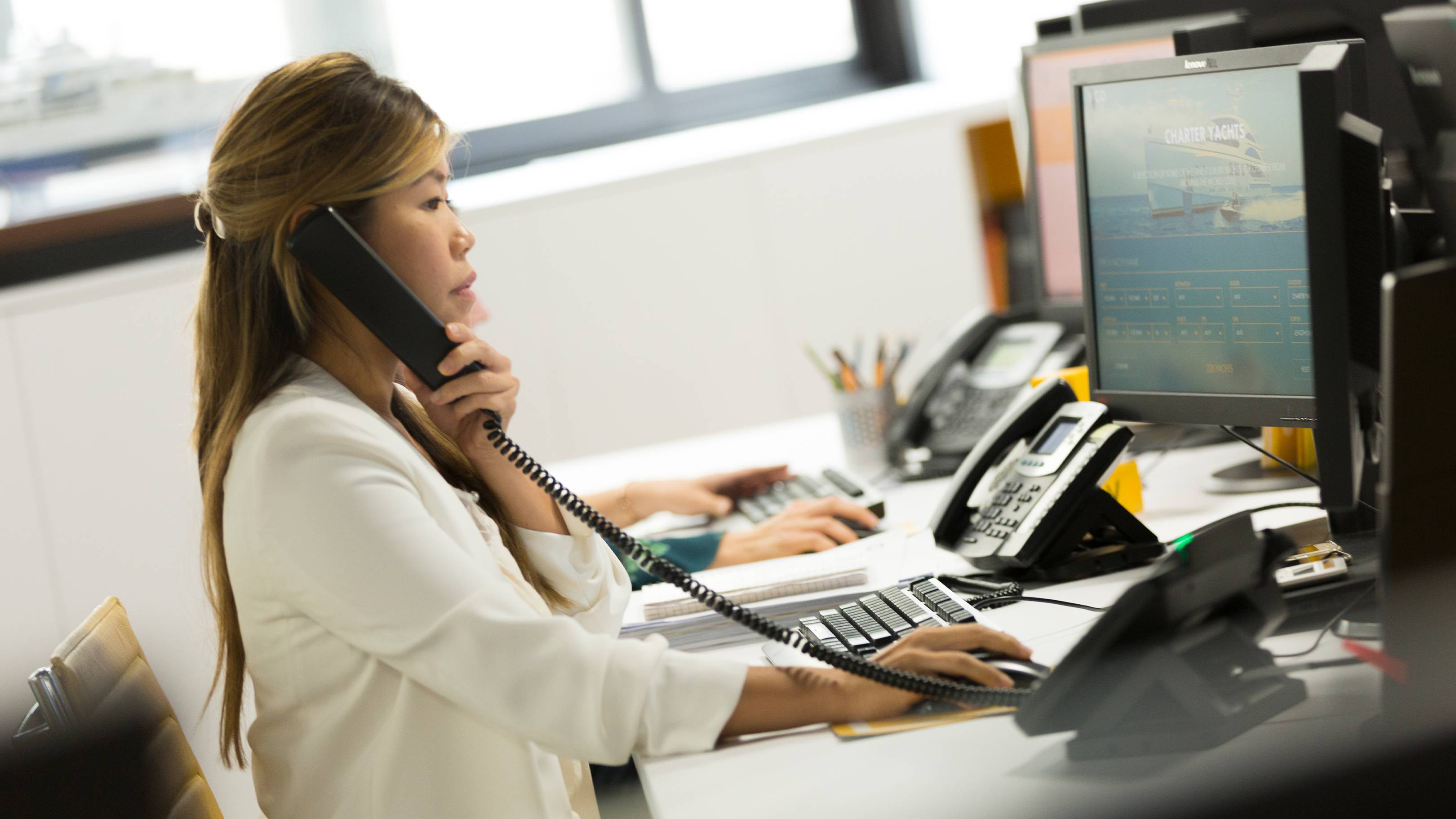 A woman in a white blouse is sitting at a desk, working on a computer and talking on a landline phone in an office setting with various office equipment and a window in the background.