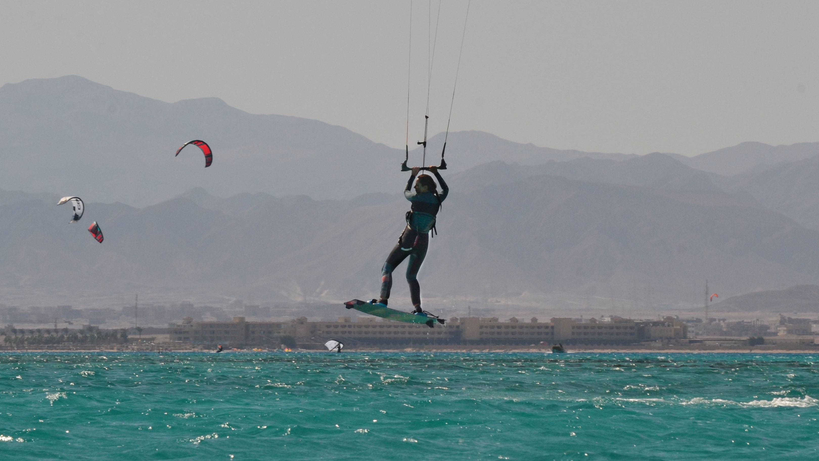 A kitesurfer soars through the air above turquoise waters with a hazy mountain range in the background.