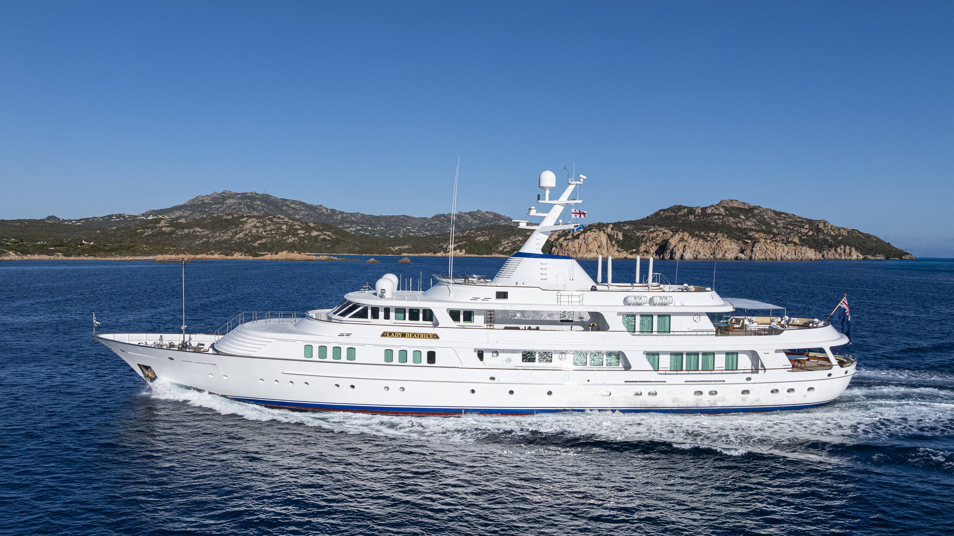 The elegant Lady Beatrice yacht cruises through the deep blue waters with a backdrop of rugged, tree-covered islands.