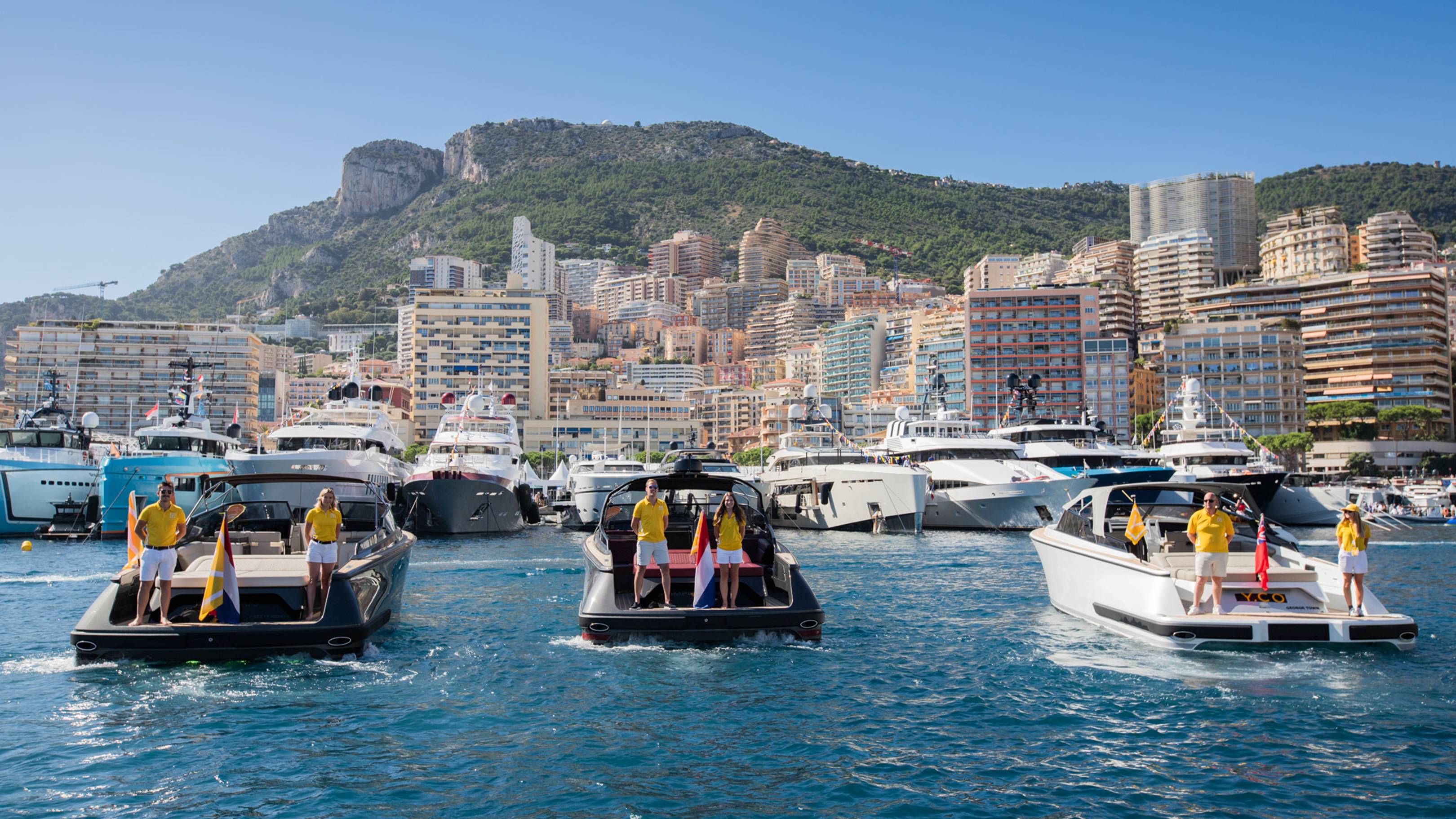 Luxury yachts cruise the azure waters of a Mediterranean harbor, with a densely populated hillside city as a backdrop.