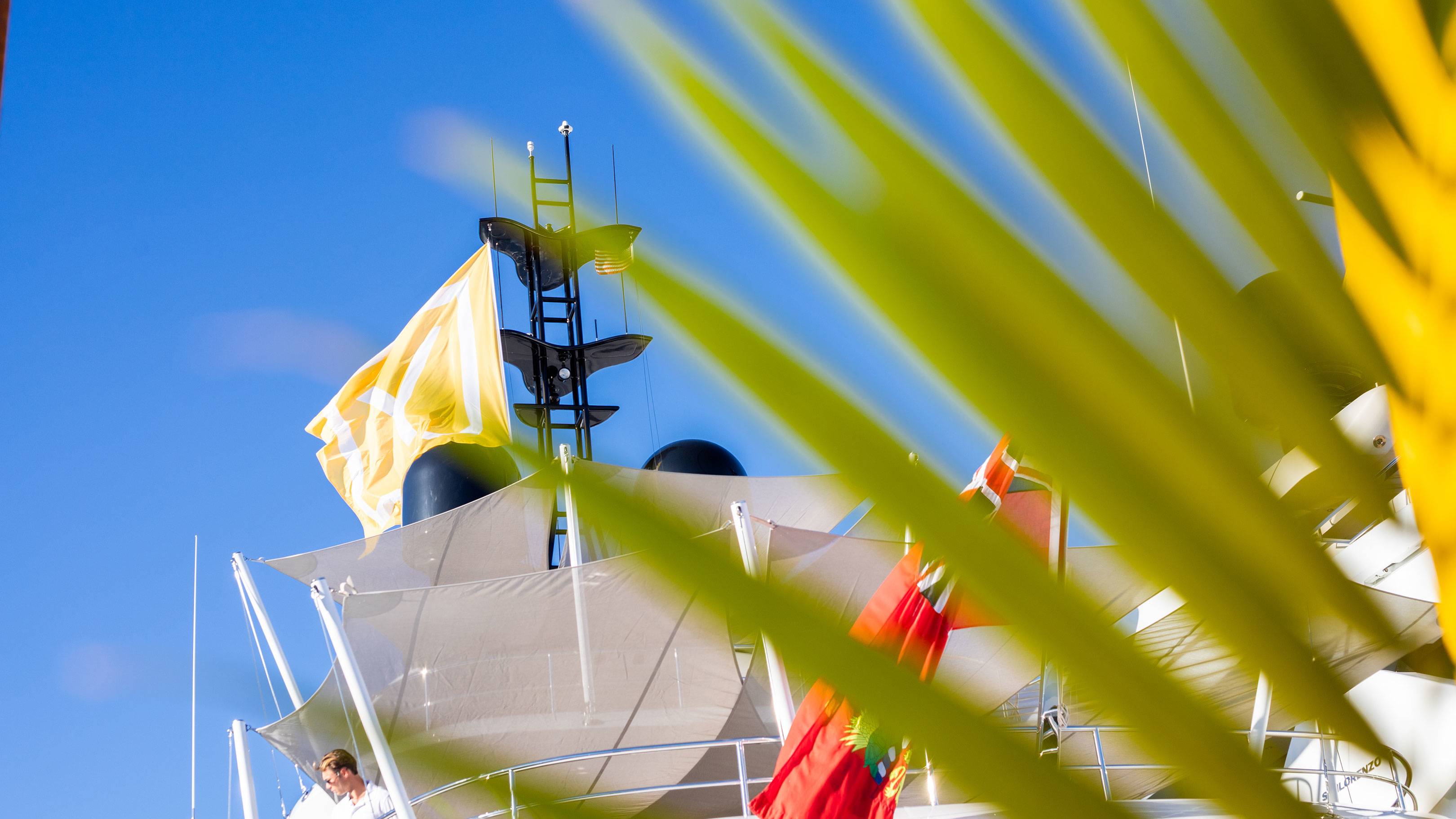 A man in a white shirt looks out from the deck of a yacht flying colorful flags under a bright blue sky.