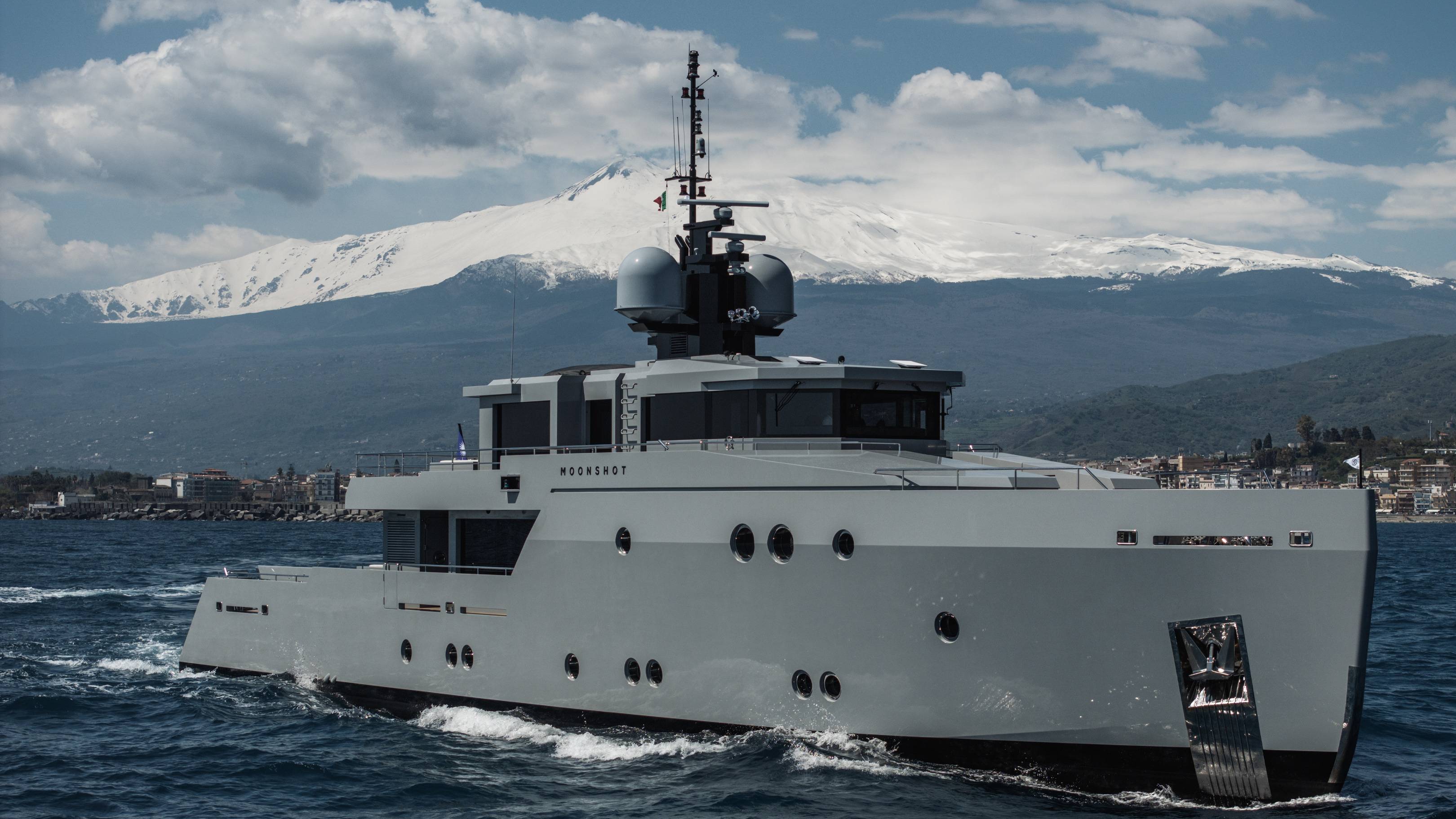 A large, sleek white yacht navigates the calm waters, with snow-capped mountains visible in the background against a cloudy sky.