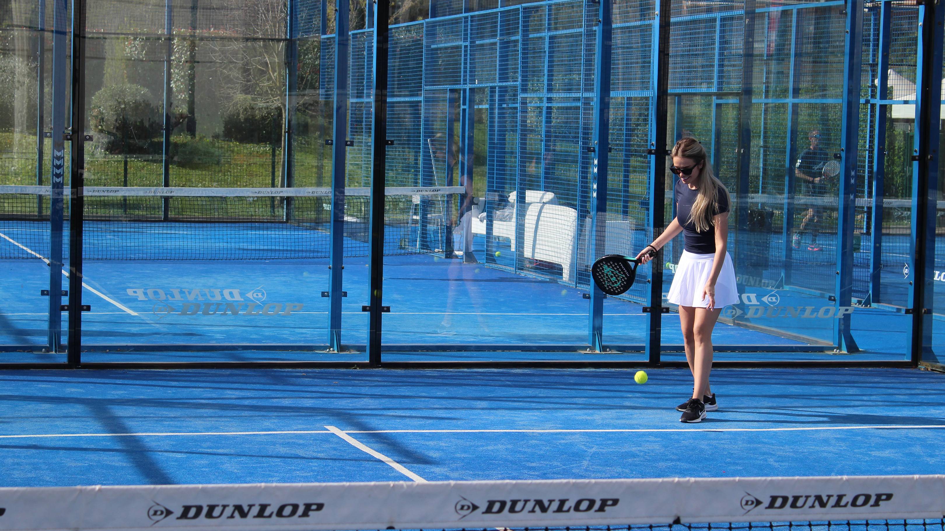 A woman in a white skirt and dark top plays padel on a blue court with a net in the foreground and a fence and trees in the background.