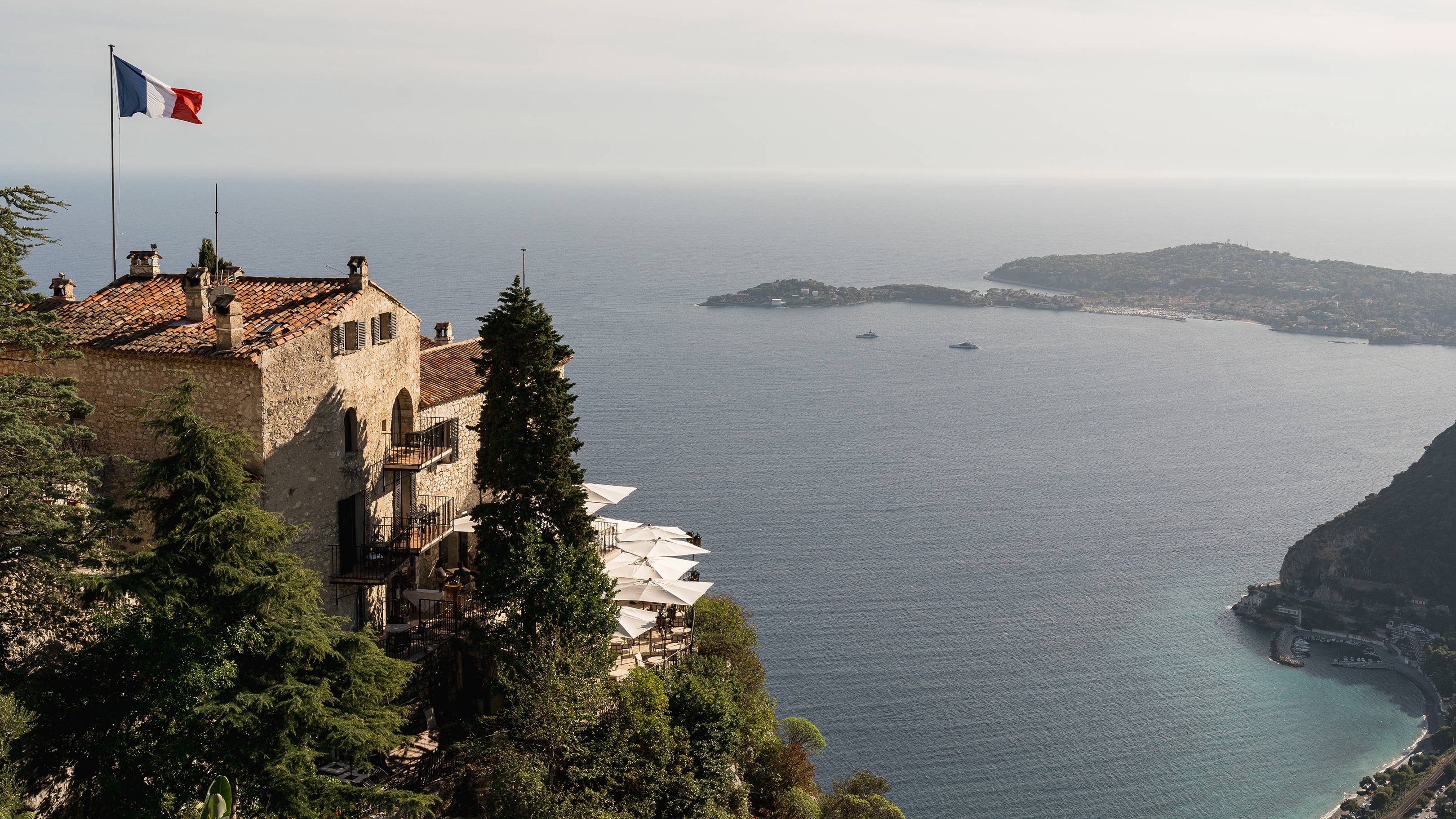 A stone building with a French flag flies on a cliff overlooking a calm blue sea with islands in the distance.