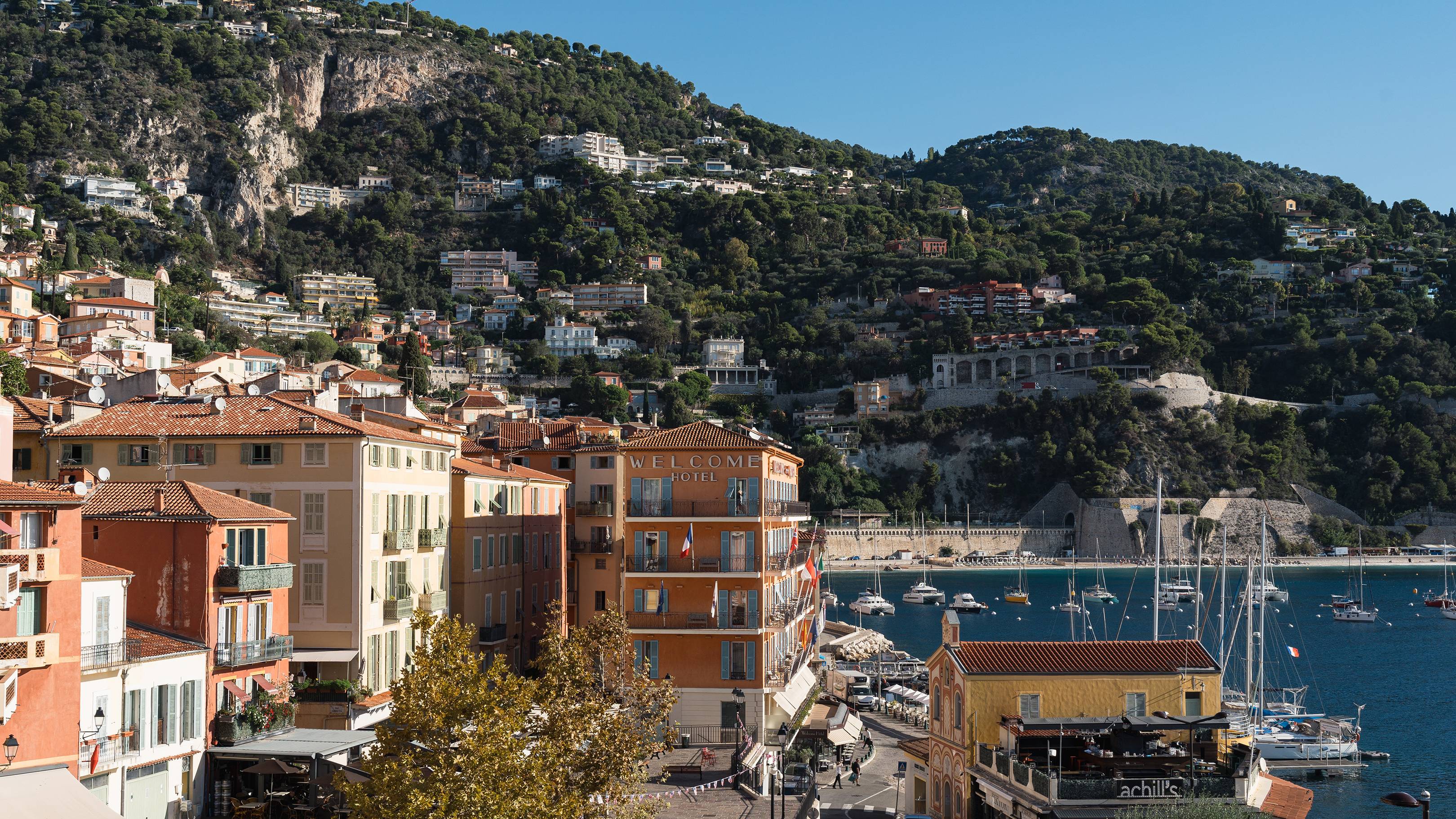Colorful buildings line a waterfront street in the foreground, with a bay dotted with sailboats and a lush, green hillside dotted with more buildings in the background.