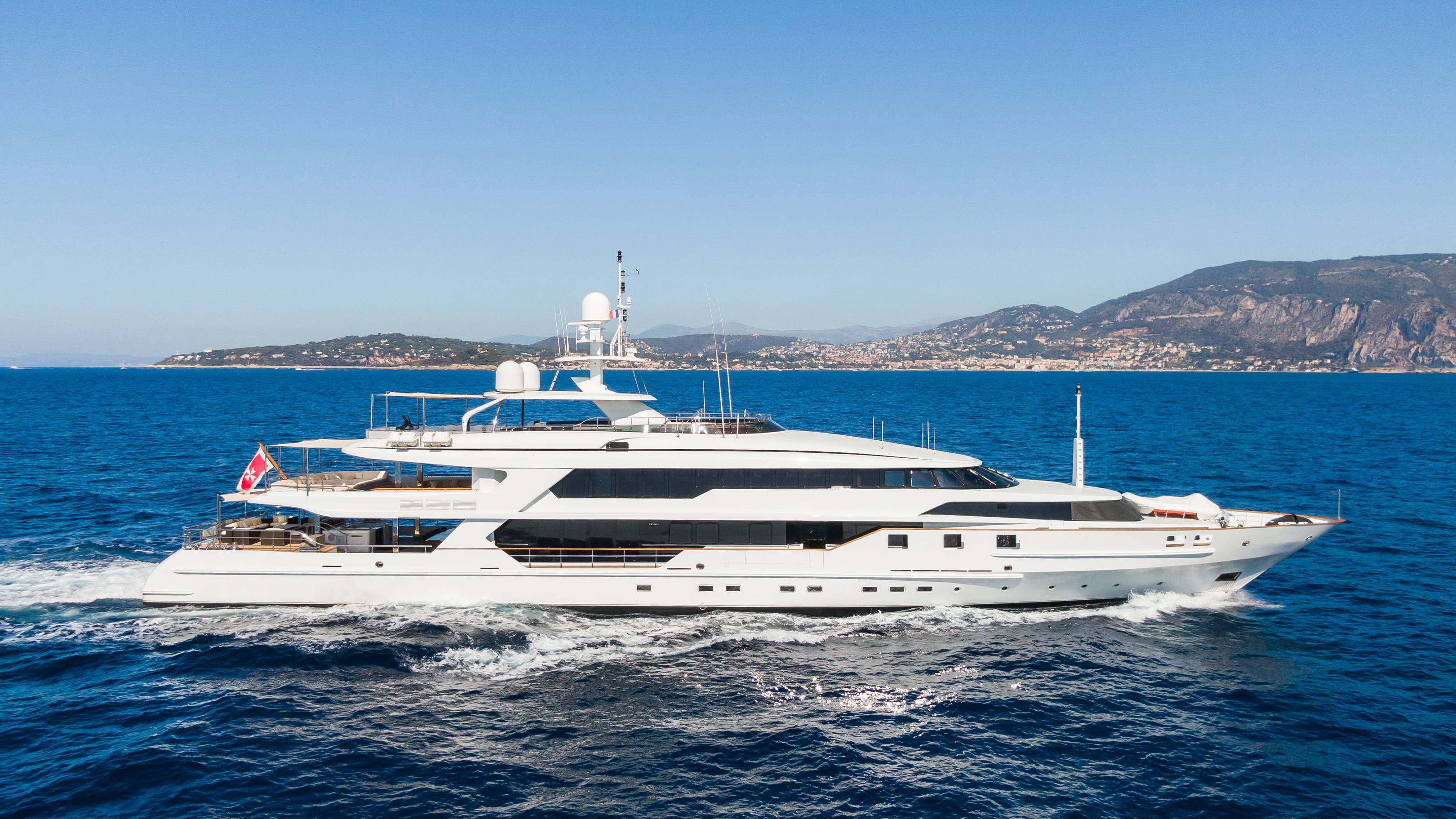 A large white yacht cruises through the blue ocean with a hilly coastline in the background.