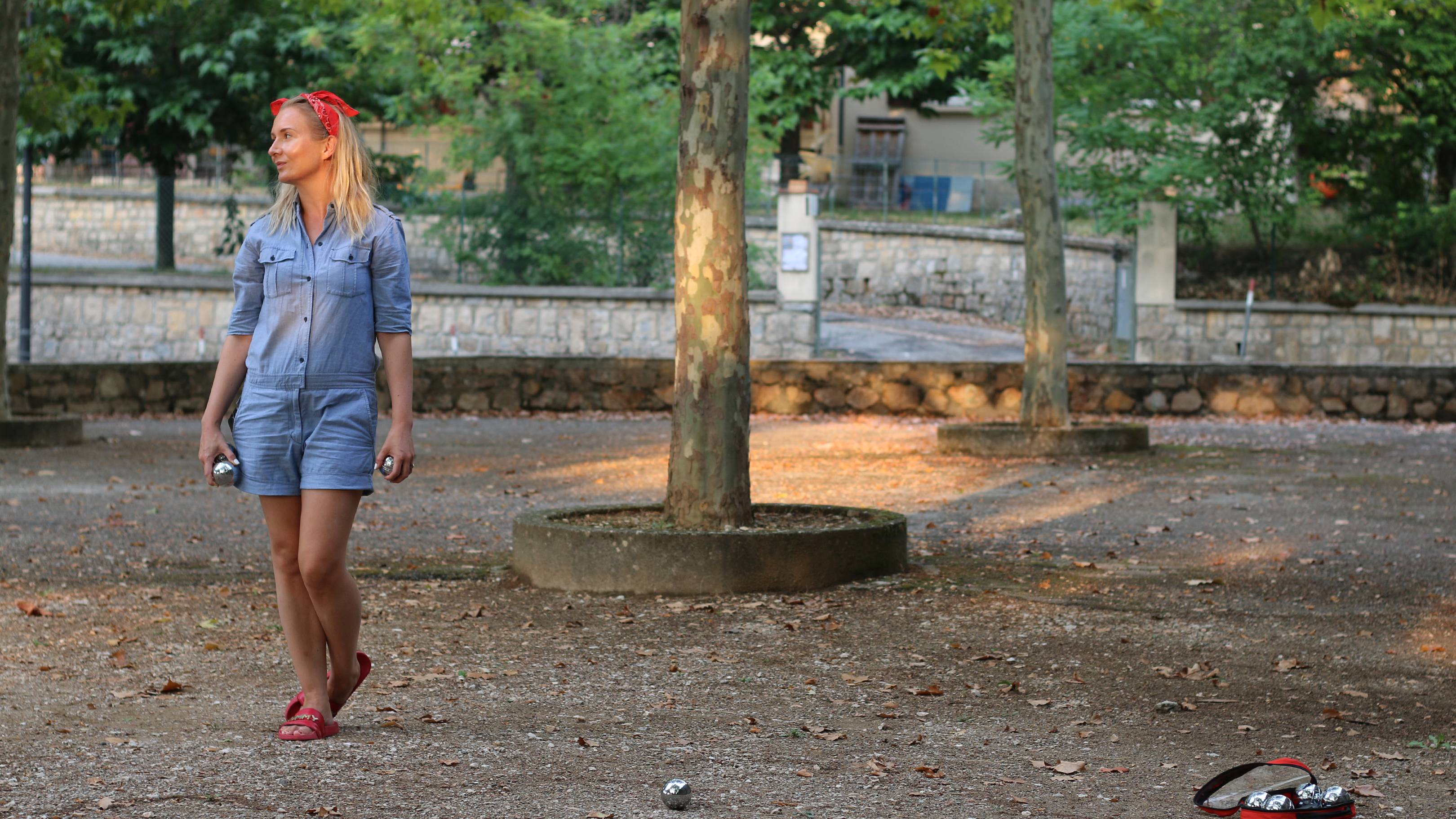 A woman in a denim romper stands in a park with petanque balls scattered on the ground, while trees and a stone wall form the background.