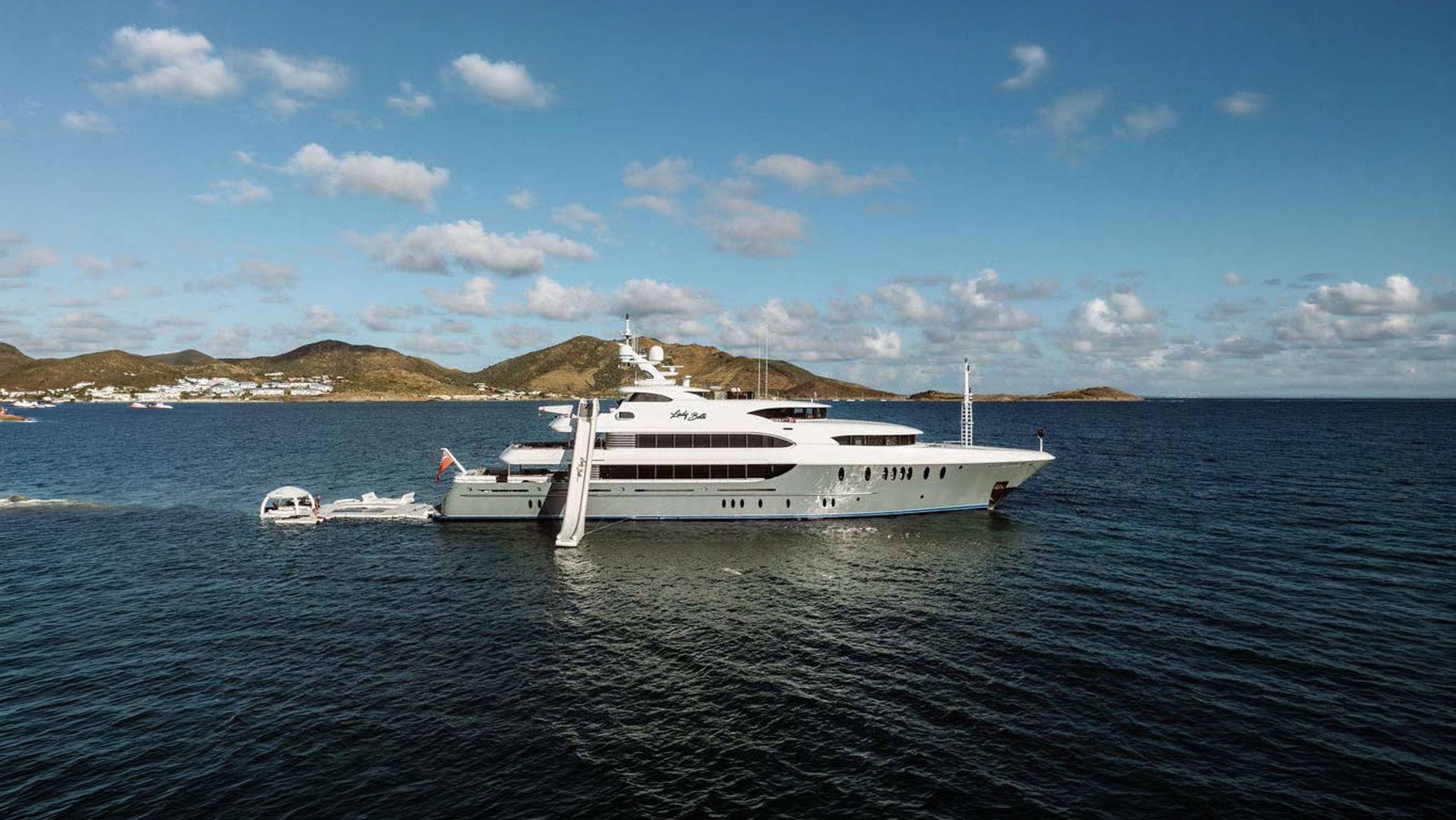 A large white yacht with a slide is anchored in the deep blue sea, with a hilly coastline dotted with white buildings in the background under a cloudy sky.