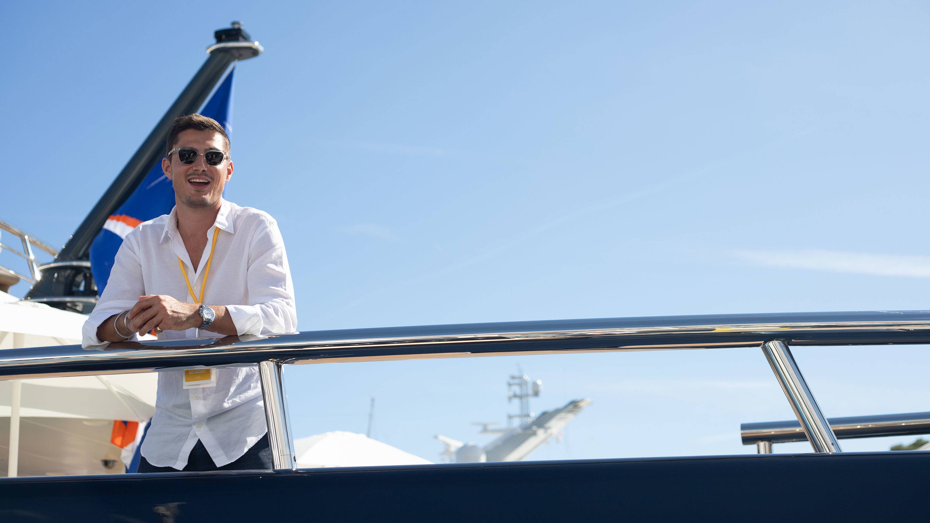 A man in a white shirt smiles on a yacht's deck with a clear blue sky and another boat in the background.