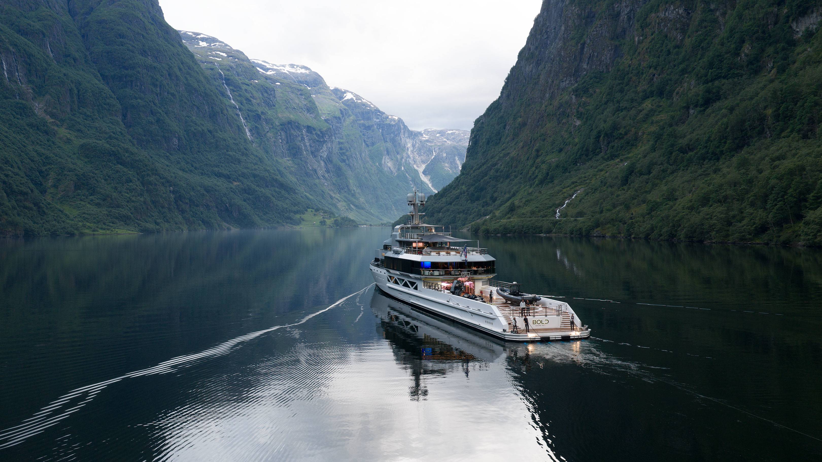 A luxurious yacht cruises through a calm, reflective fjord, flanked by steep, verdant mountains under a cloudy sky.