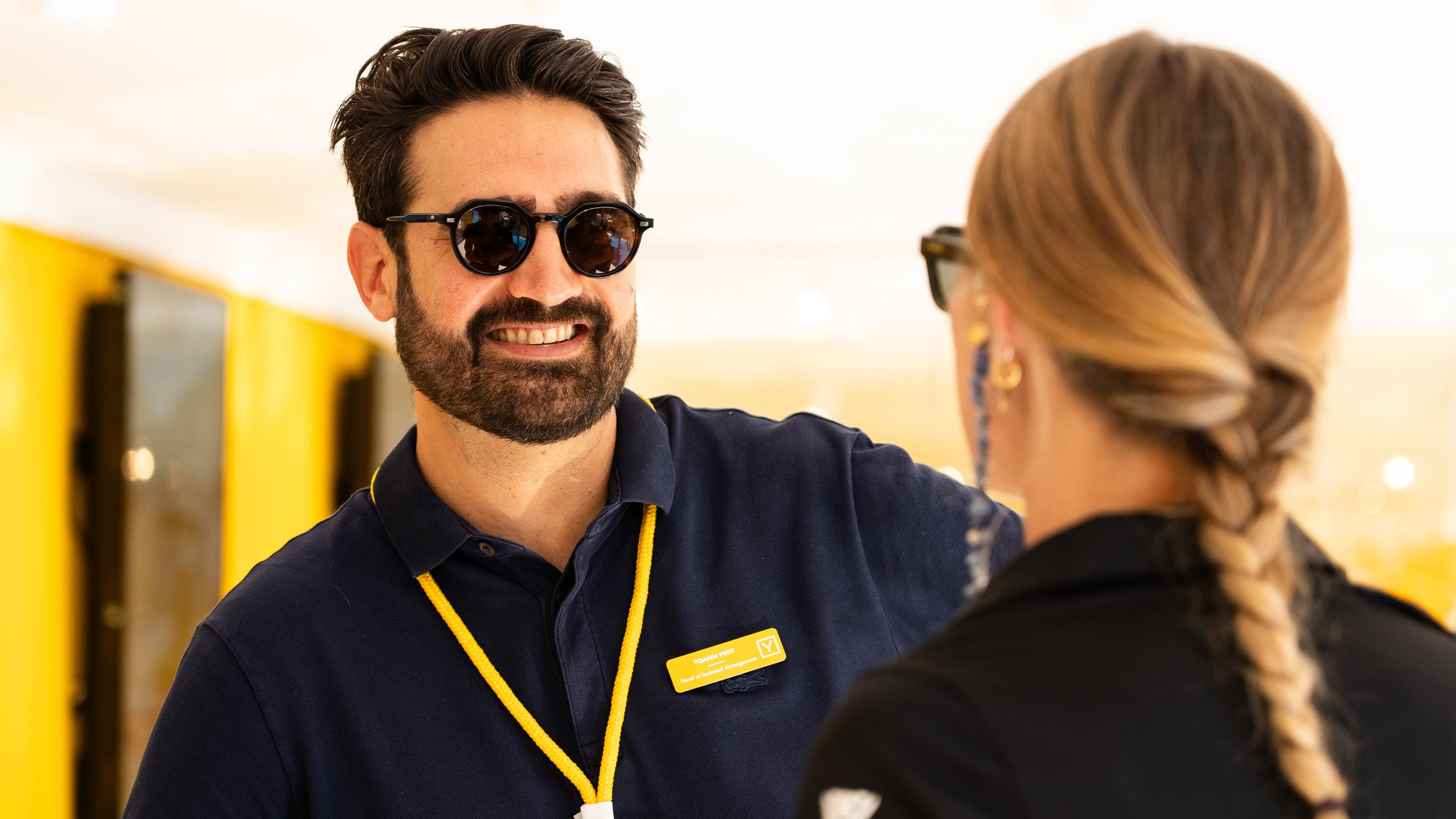 A smiling man in sunglasses and a blue polo shirt talks to a woman with a braided blonde ponytail in the foreground, while a blurred yellow and black background is visible behind them.