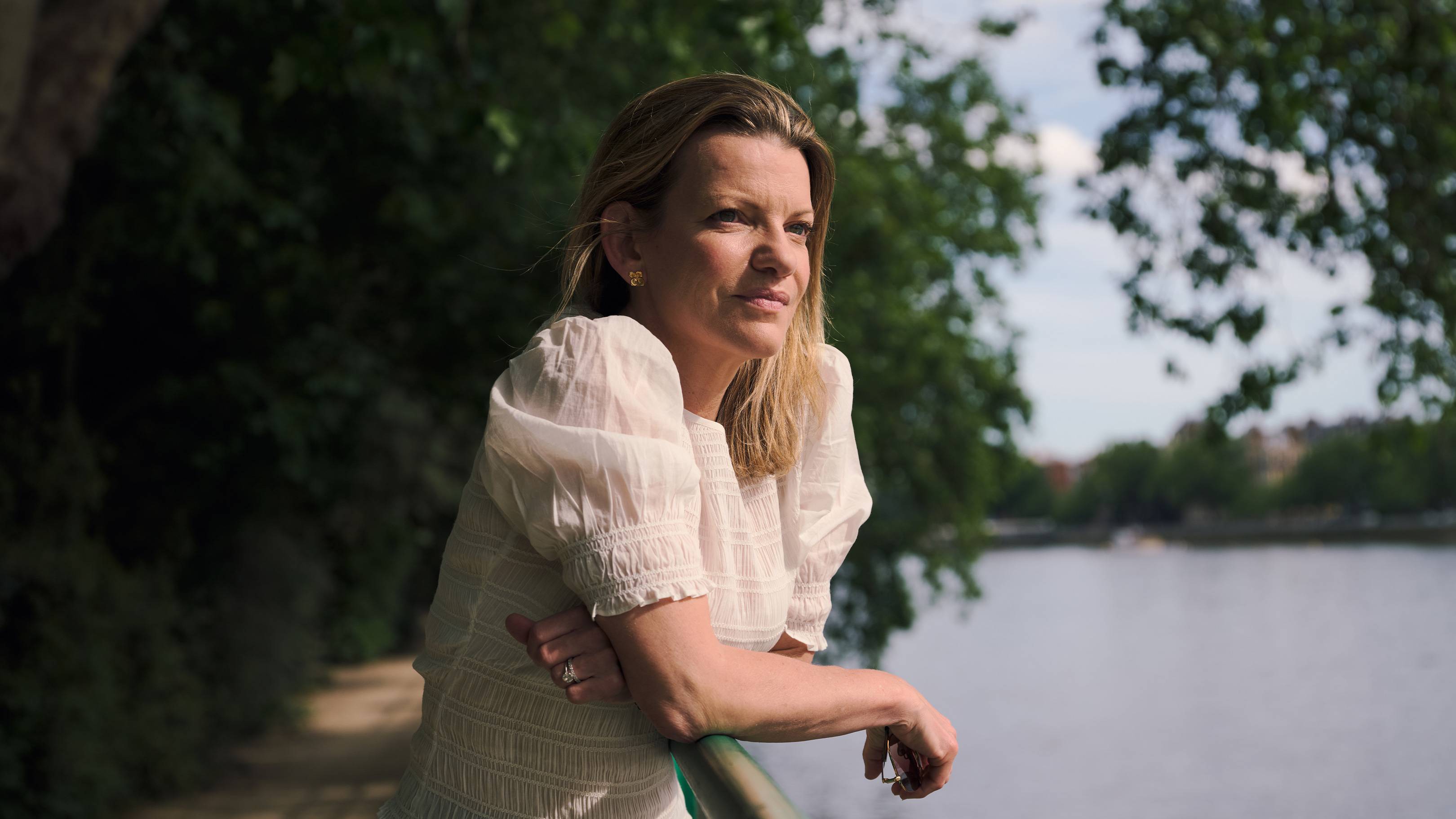 A woman in a white blouse leans on a railing by a path, looking out at a calm river with trees in the background.