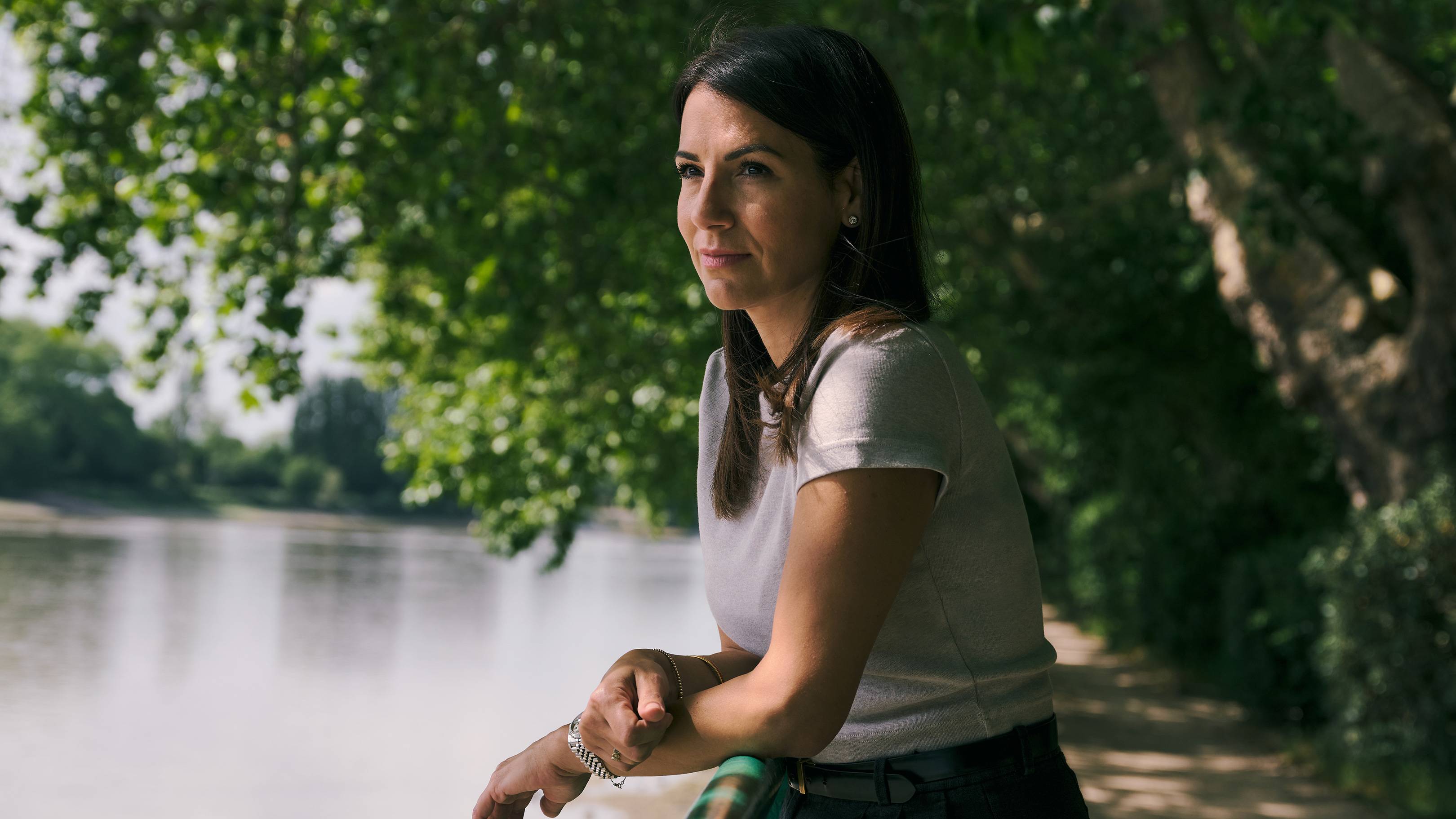 A woman leans on a railing in the foreground, looking out at a calm body of water bordered by trees in the background.