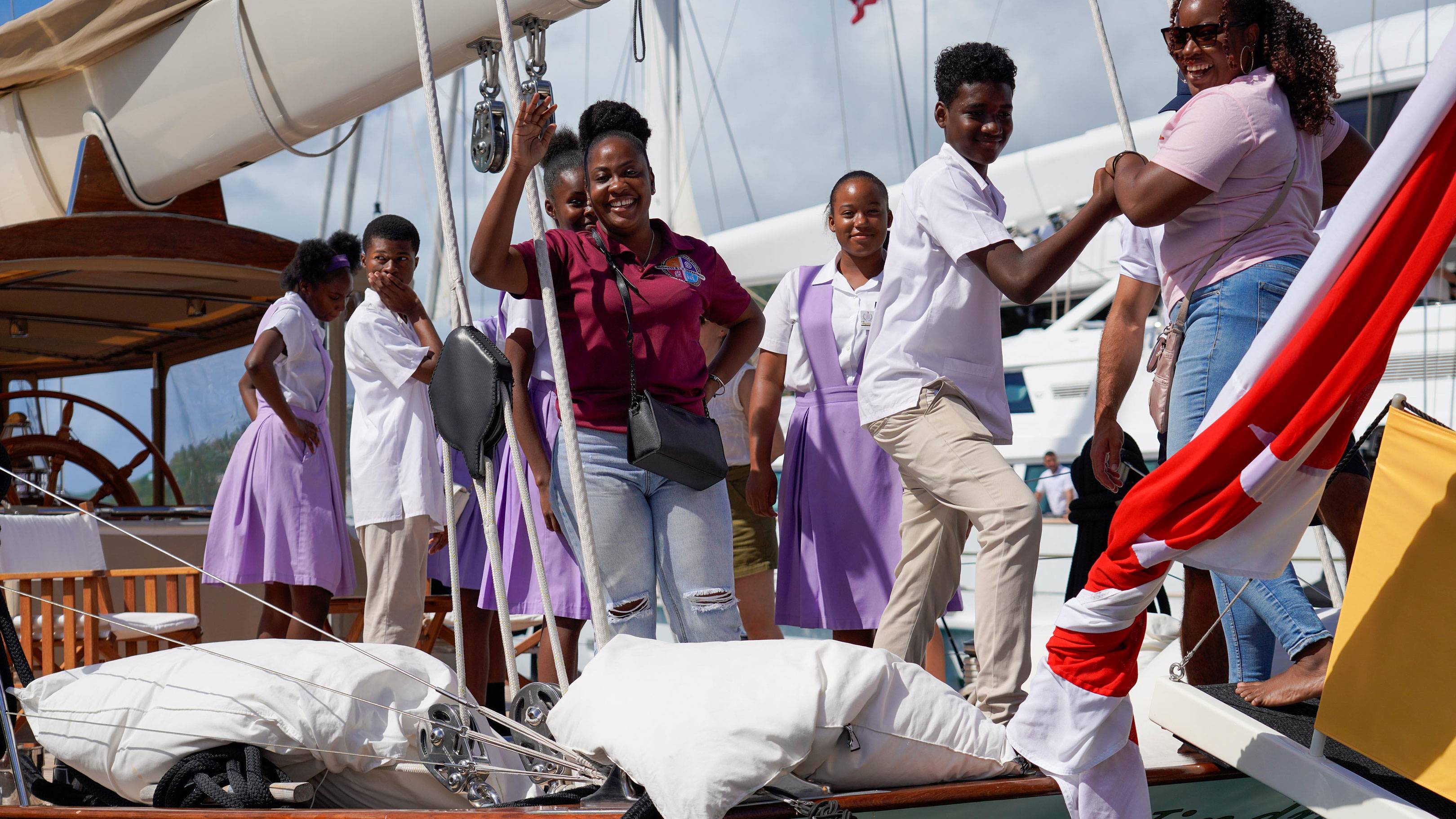 A group of smiling people, some in uniform, stand on a sailboat in the foreground, with a larger white yacht visible in the background.