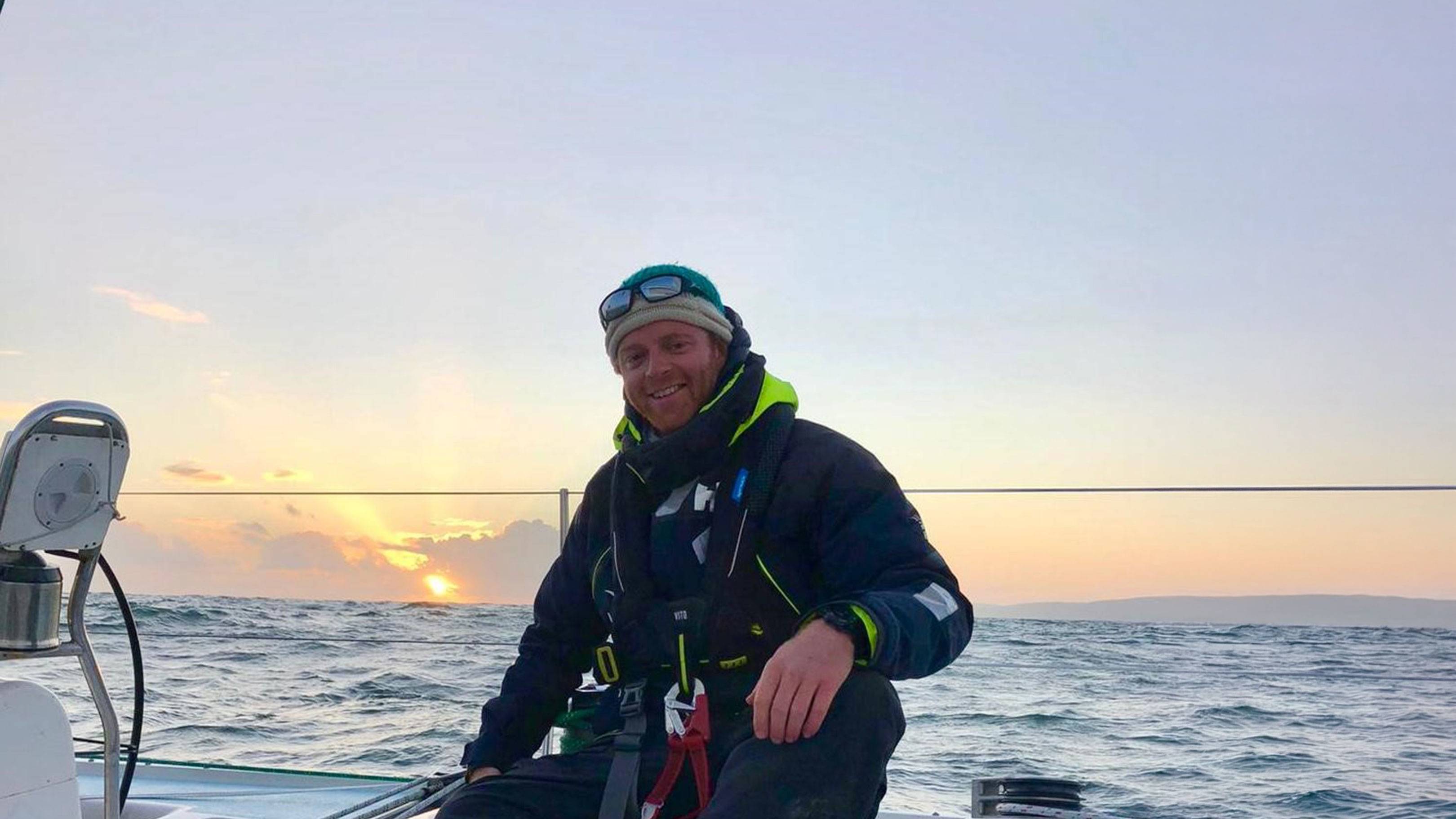 A man in sailing gear smiles on a boat's deck with a sunset over the ocean in the background.