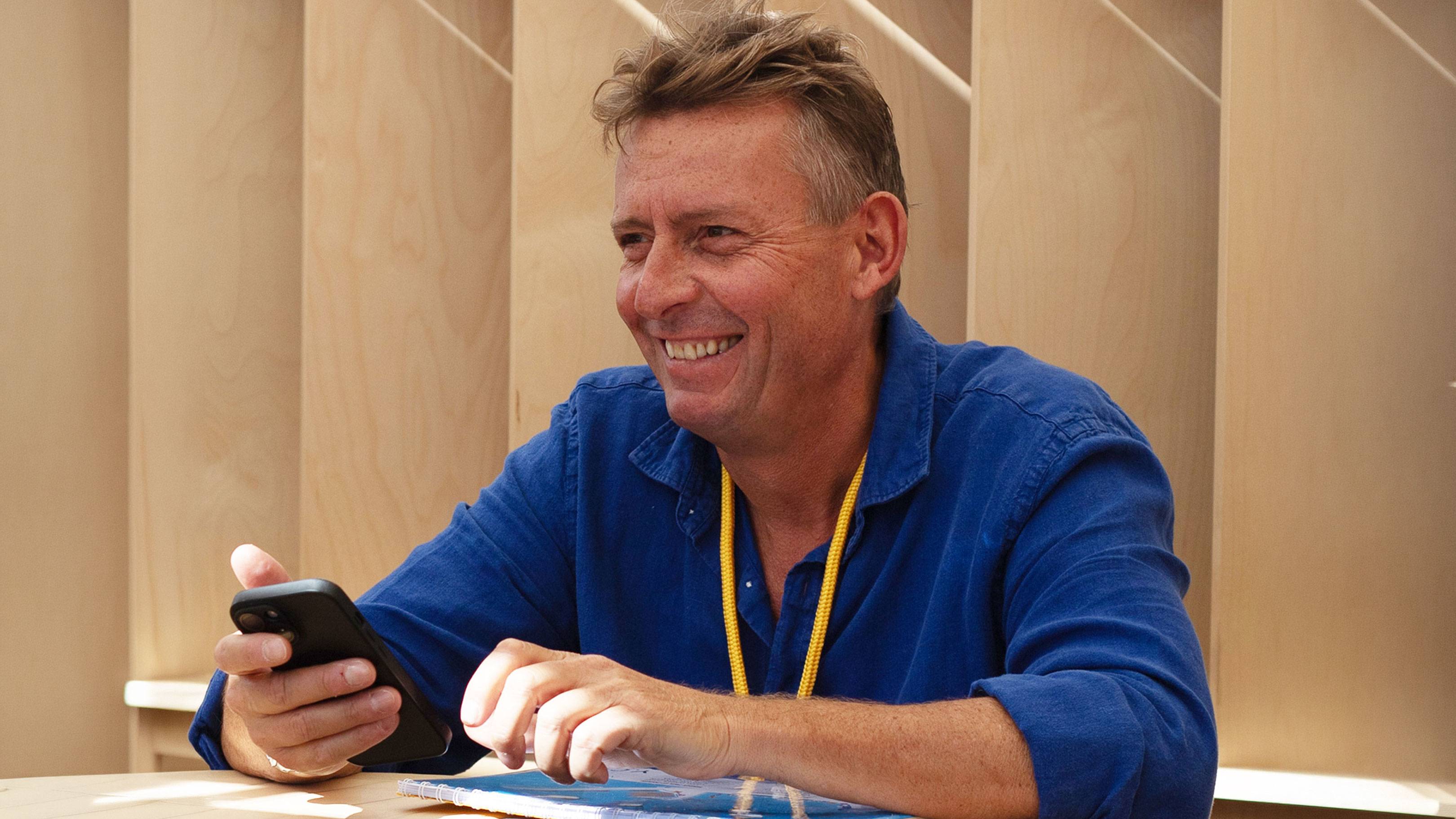 A smiling man in a blue shirt holds a smartphone in the foreground, set against a background of light-colored wooden panels.