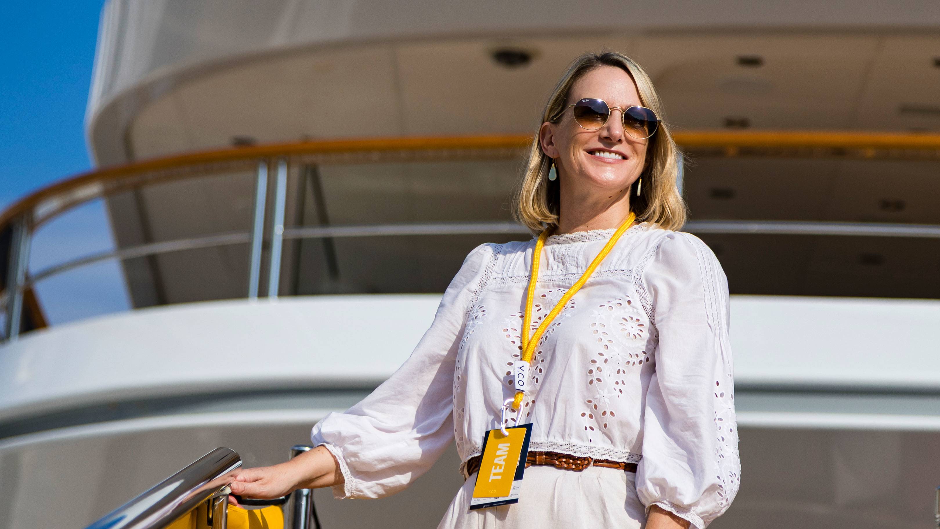 A smiling woman wearing sunglasses and a white eyelet blouse stands in the foreground against the backdrop of a large white yacht under a clear blue sky.