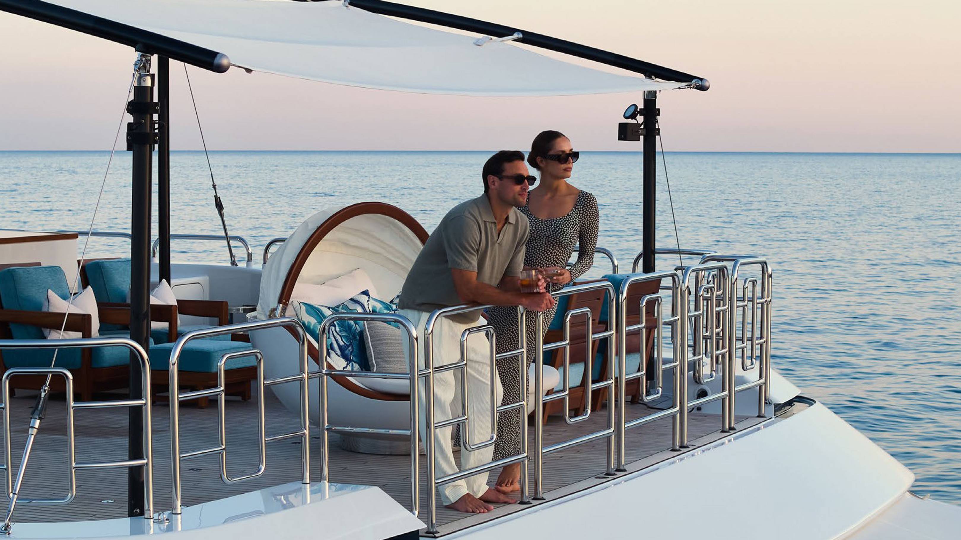 A man and woman stand on the deck of a luxury yacht, overlooking a calm blue ocean under a soft evening sky.