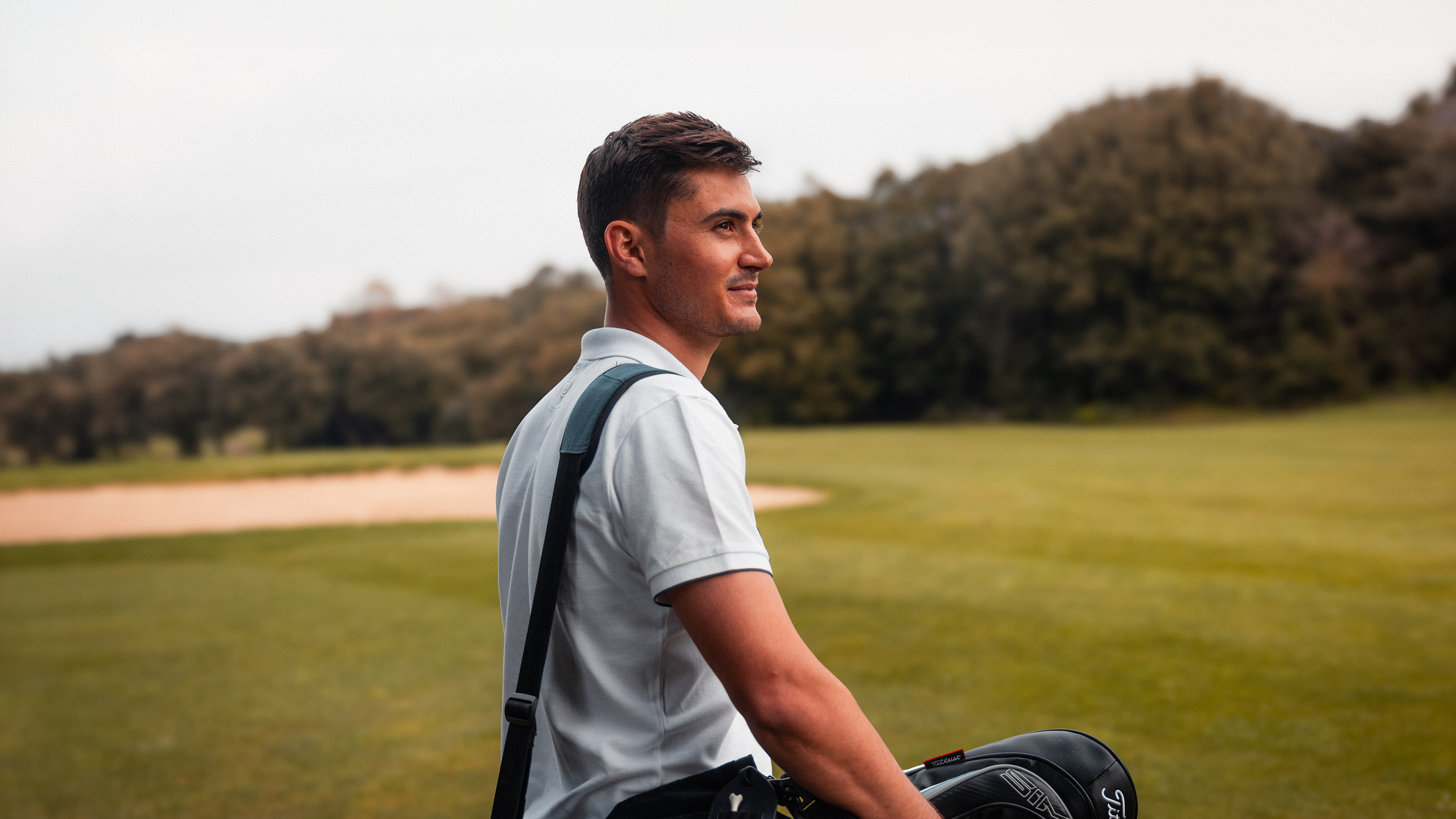A man in a white polo shirt carries a golf bag across a green course with a sand trap and wooded hills in the background.